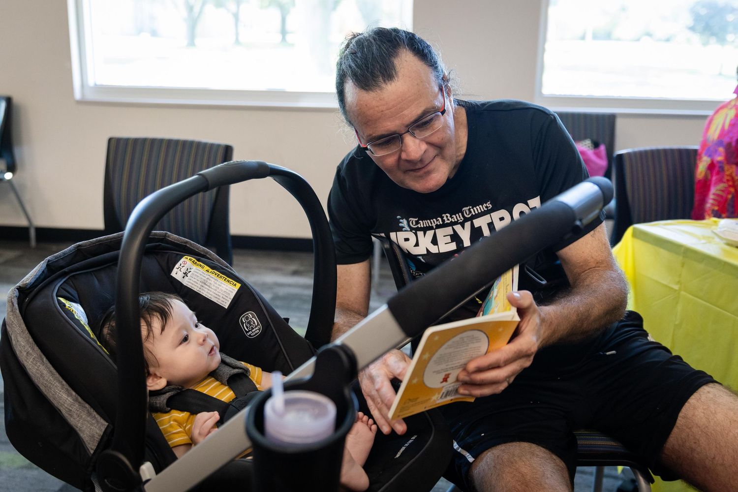 A man reads a colorful book to a baby in a car seat during a storytime session at a community center.