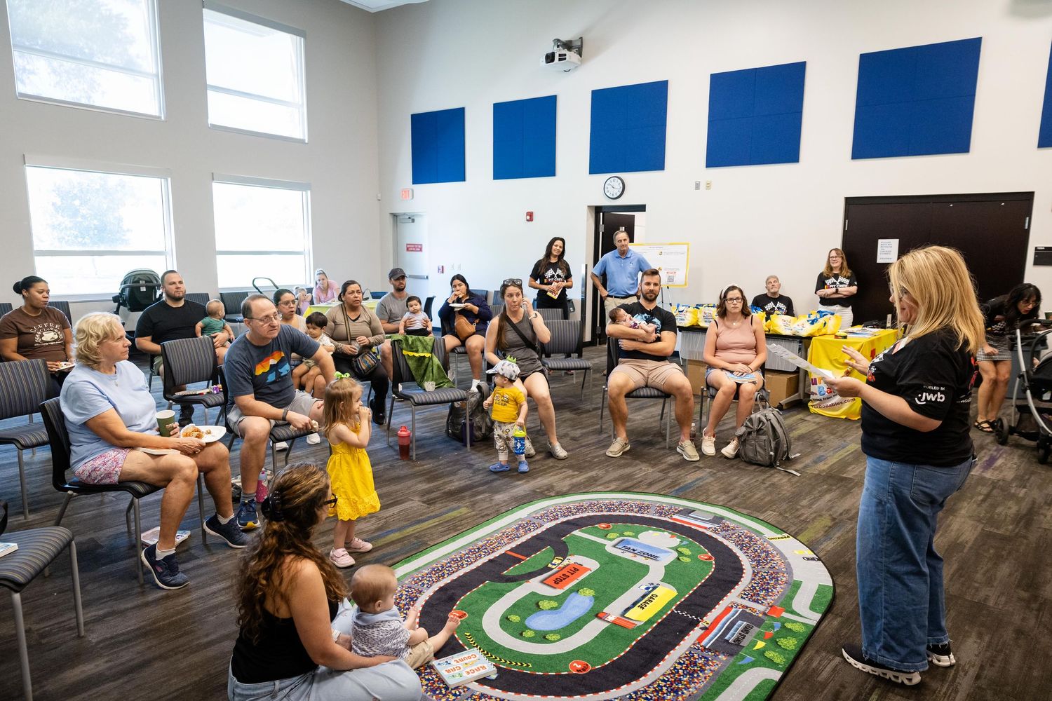 Group of families gathered in a community room listening to a presenter near a racing track floor mat.