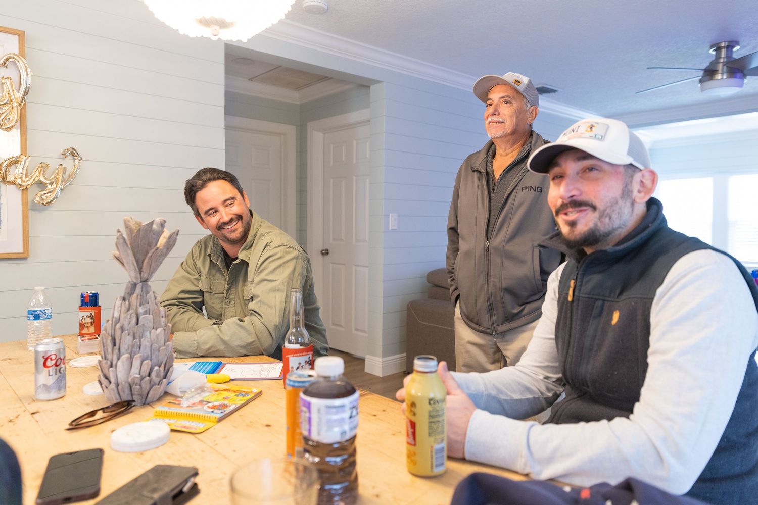 Three men smiling and relaxing around a wooden table indoors, wearing casual outdoor apparel in a cozy room.