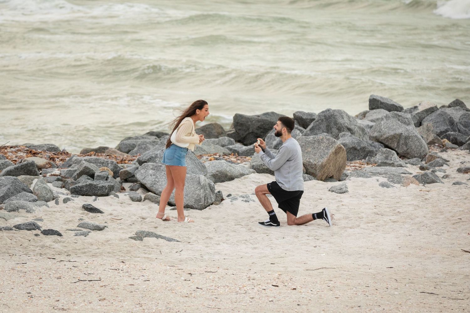 Man proposing on one knee to woman on rocky beach with ocean waves in background.