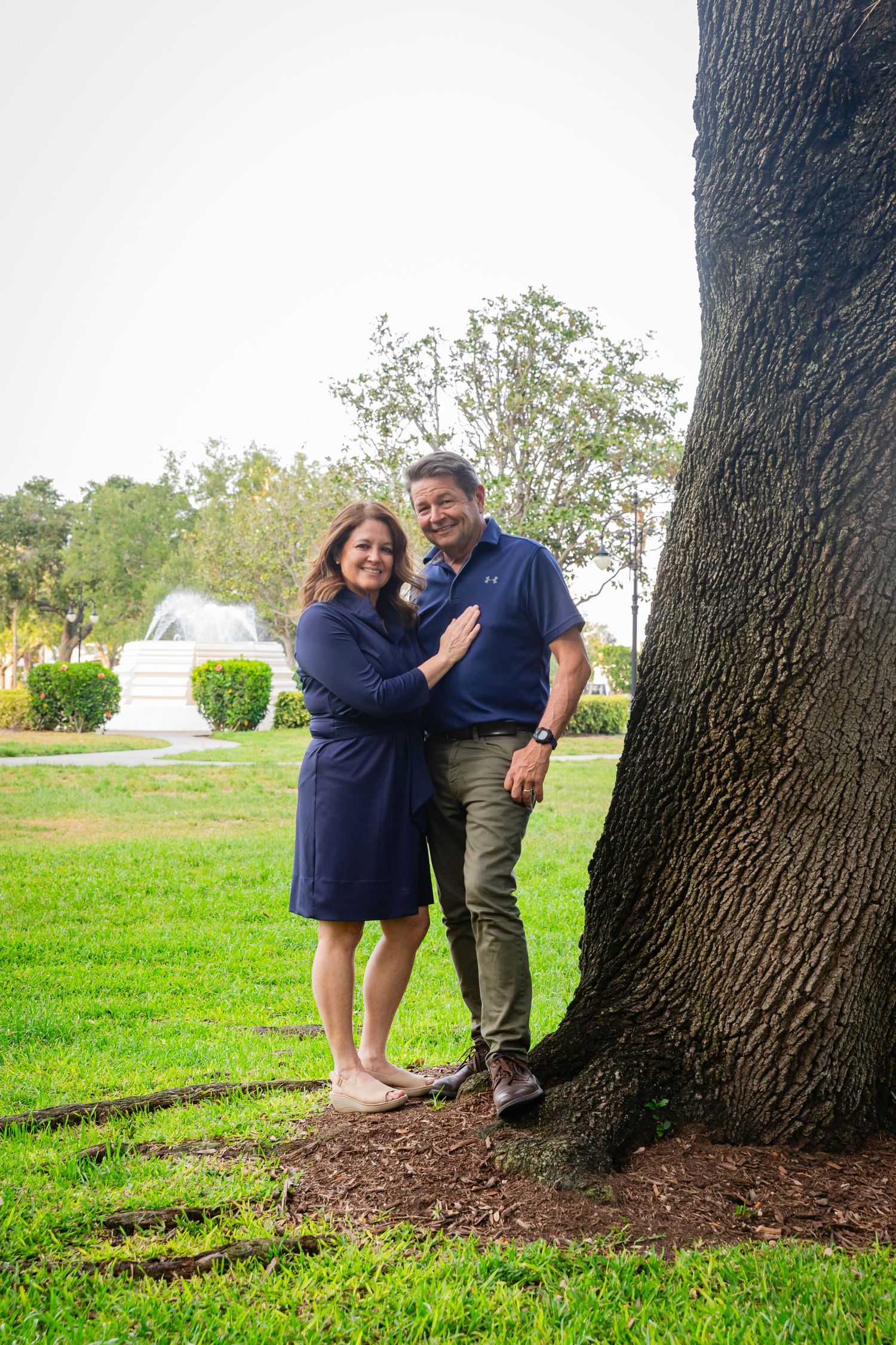 Couple in navy outfits leaning against a large tree in a lush green park with a fountain backdrop.