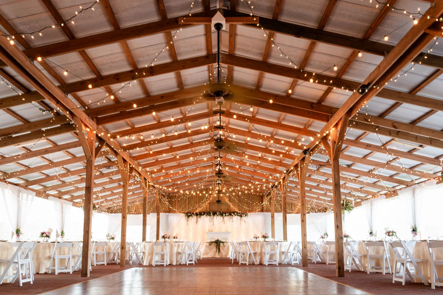 Elegant barn wedding reception venue with string lights, white draped tables, and rustic wooden beamed ceiling.