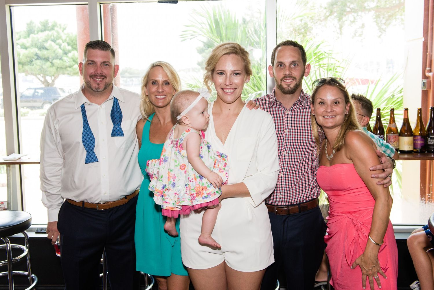 Group of six smiling adults and a baby girl posing together at a bright indoor reception venue.