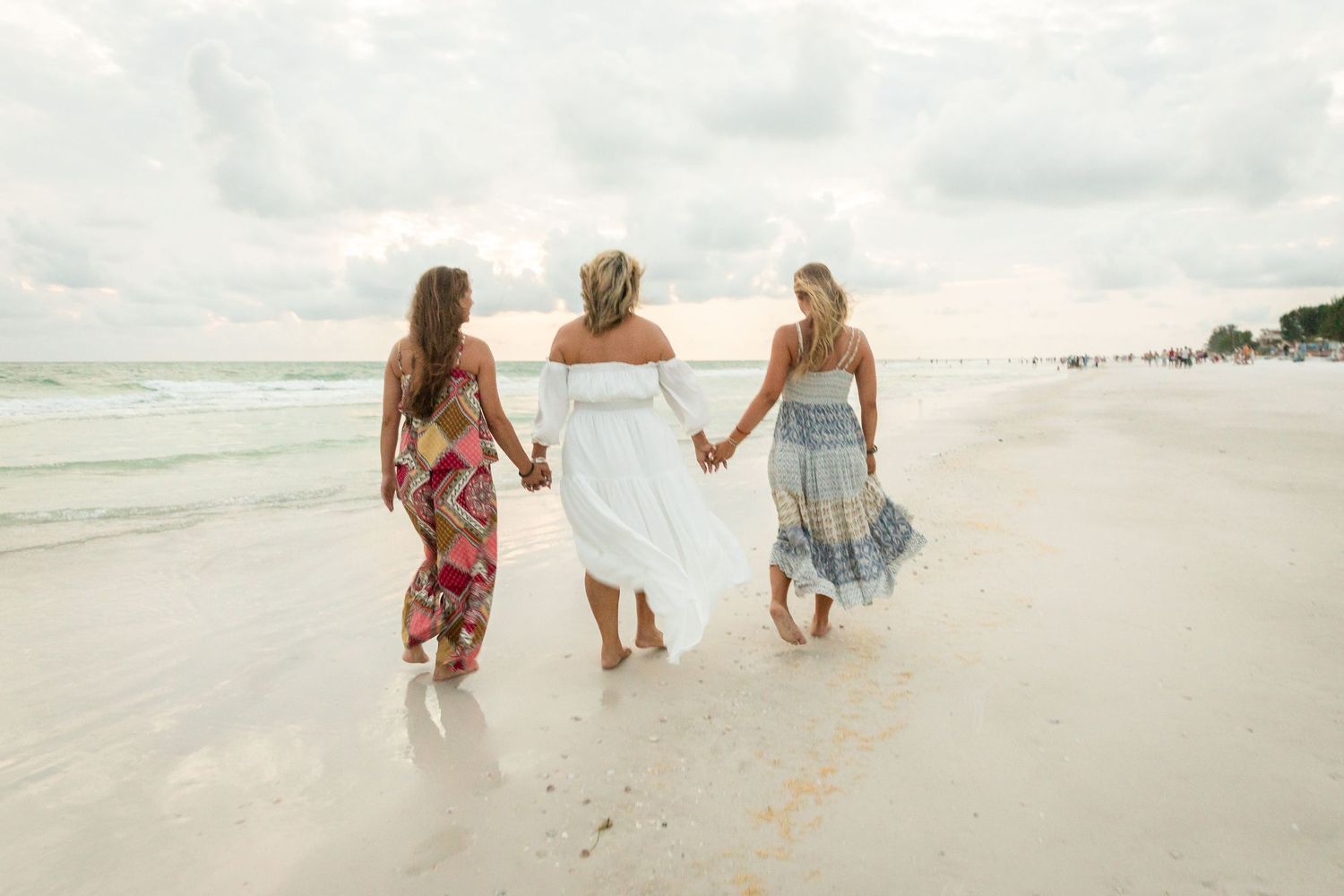 Three women in summer dresses walk hand-in-hand along a white sand beach with calm ocean waves.