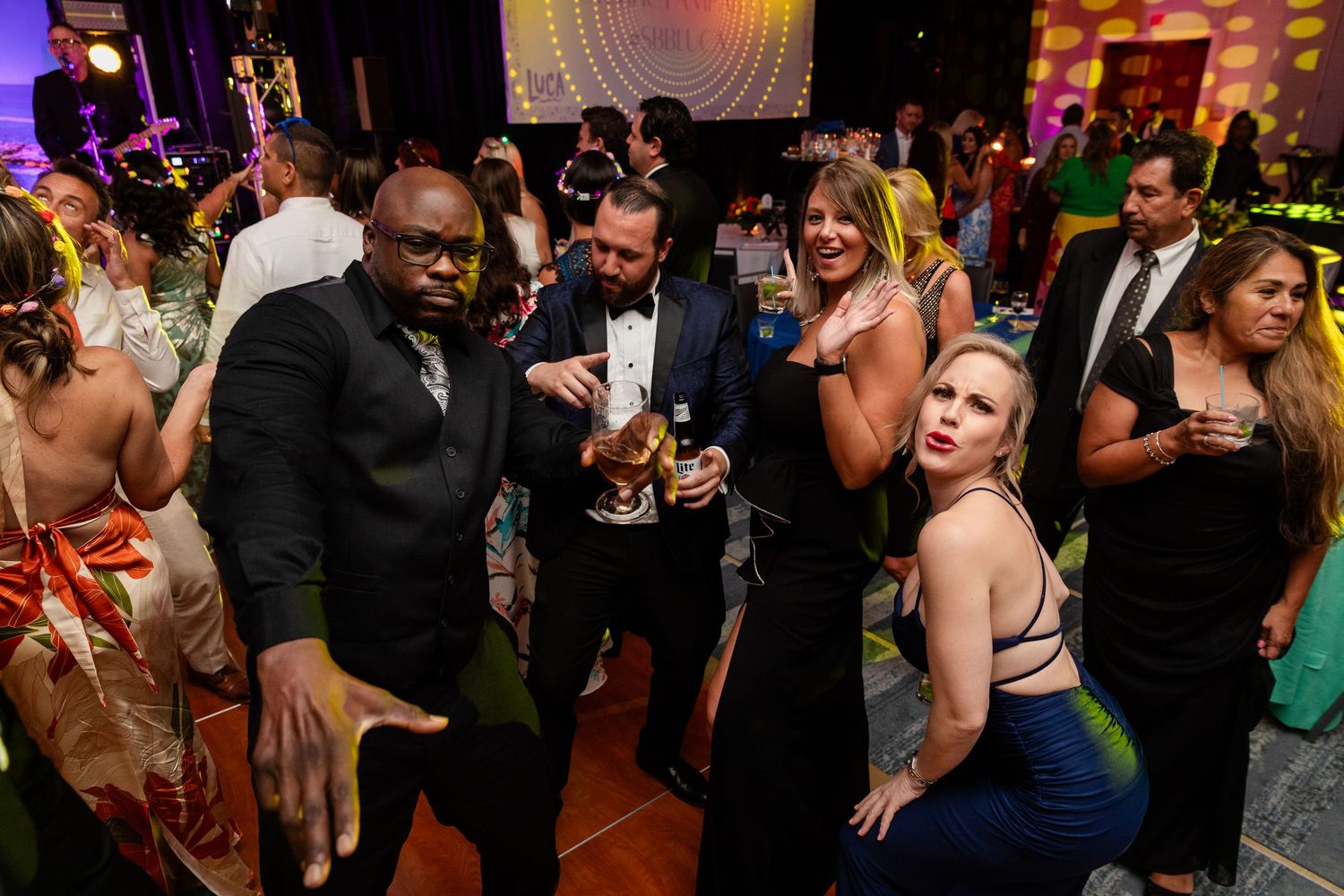 Guests in formal attire pose and dance together at an elegant evening gala event with colorful lighting.