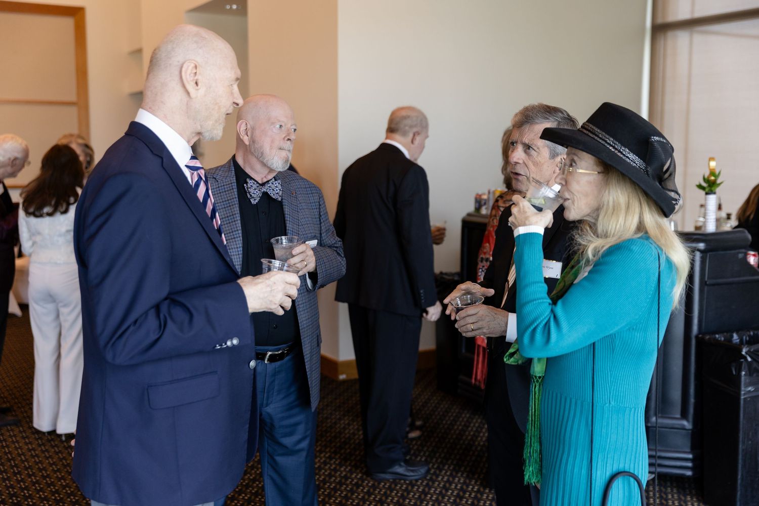 Guests networking and conversing at a formal cocktail reception, with a woman in teal and black hat socializing.