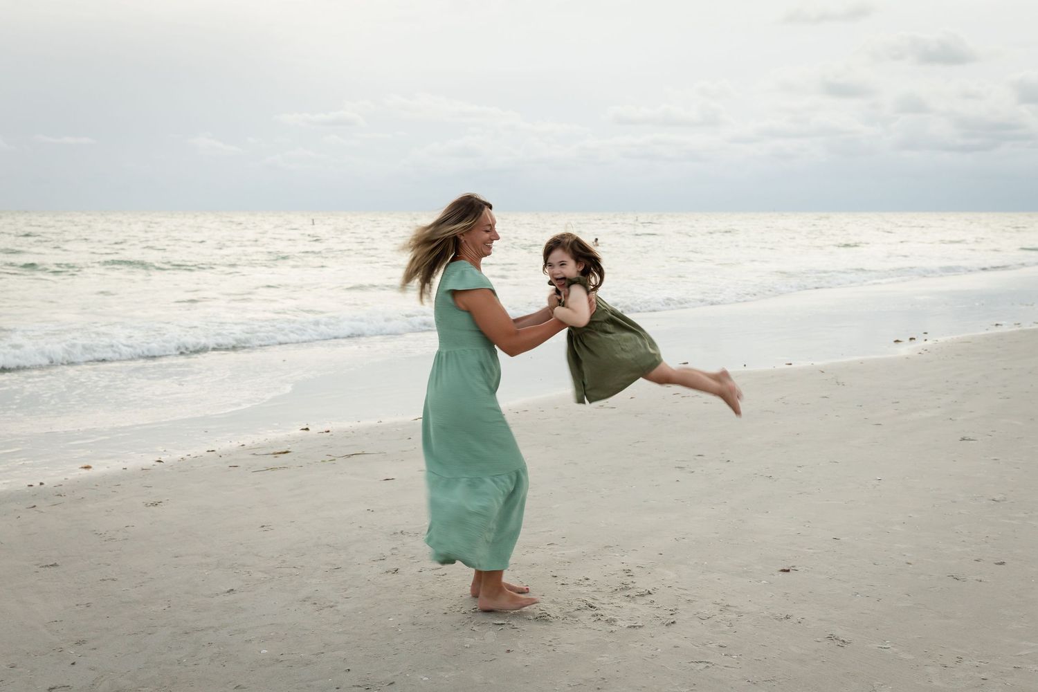 A mother in a teal dress swings her laughing child on a sandy beach with ocean waves in the background.