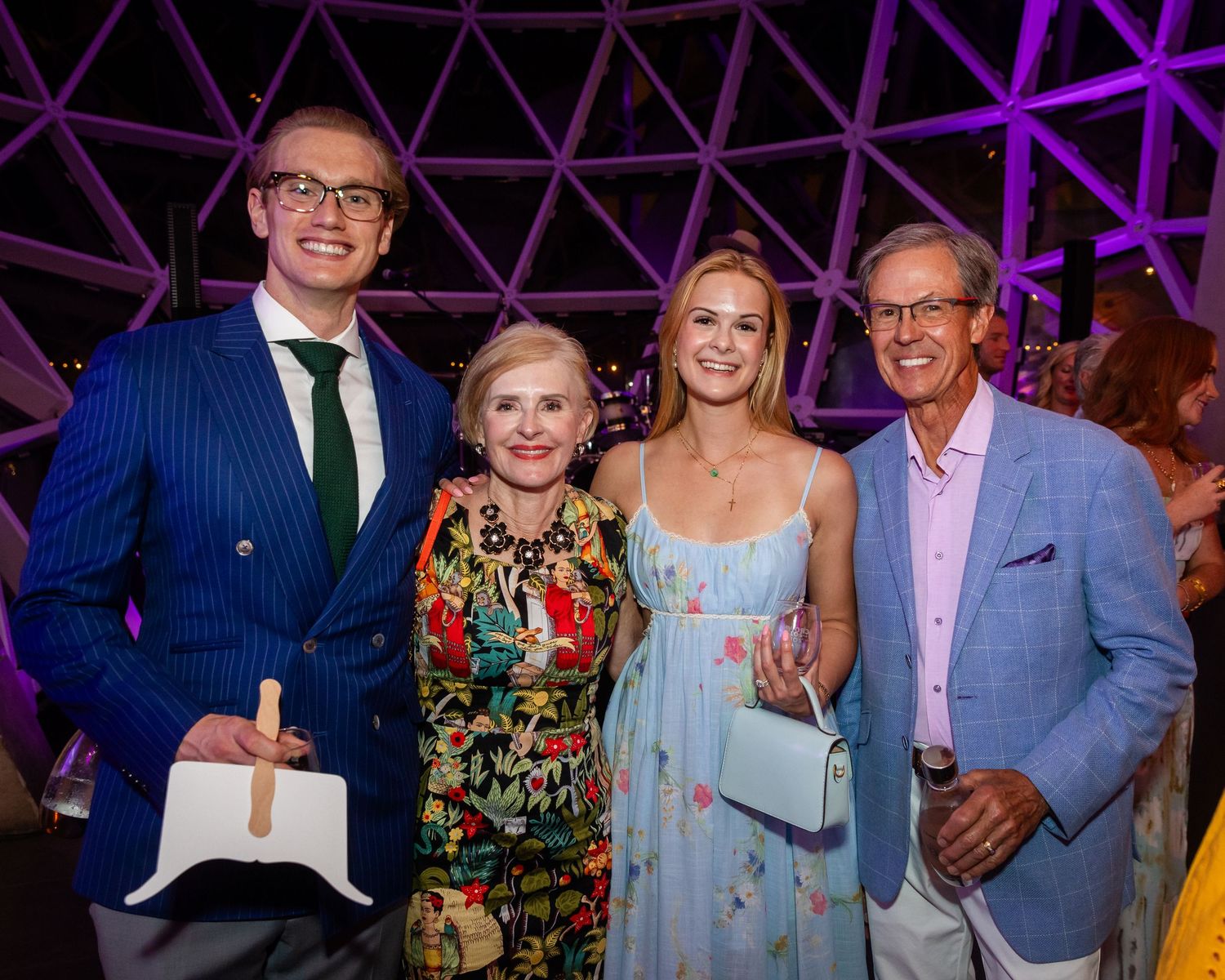 Four elegantly dressed guests smiling together at a glamorous evening gala event with purple lighting.