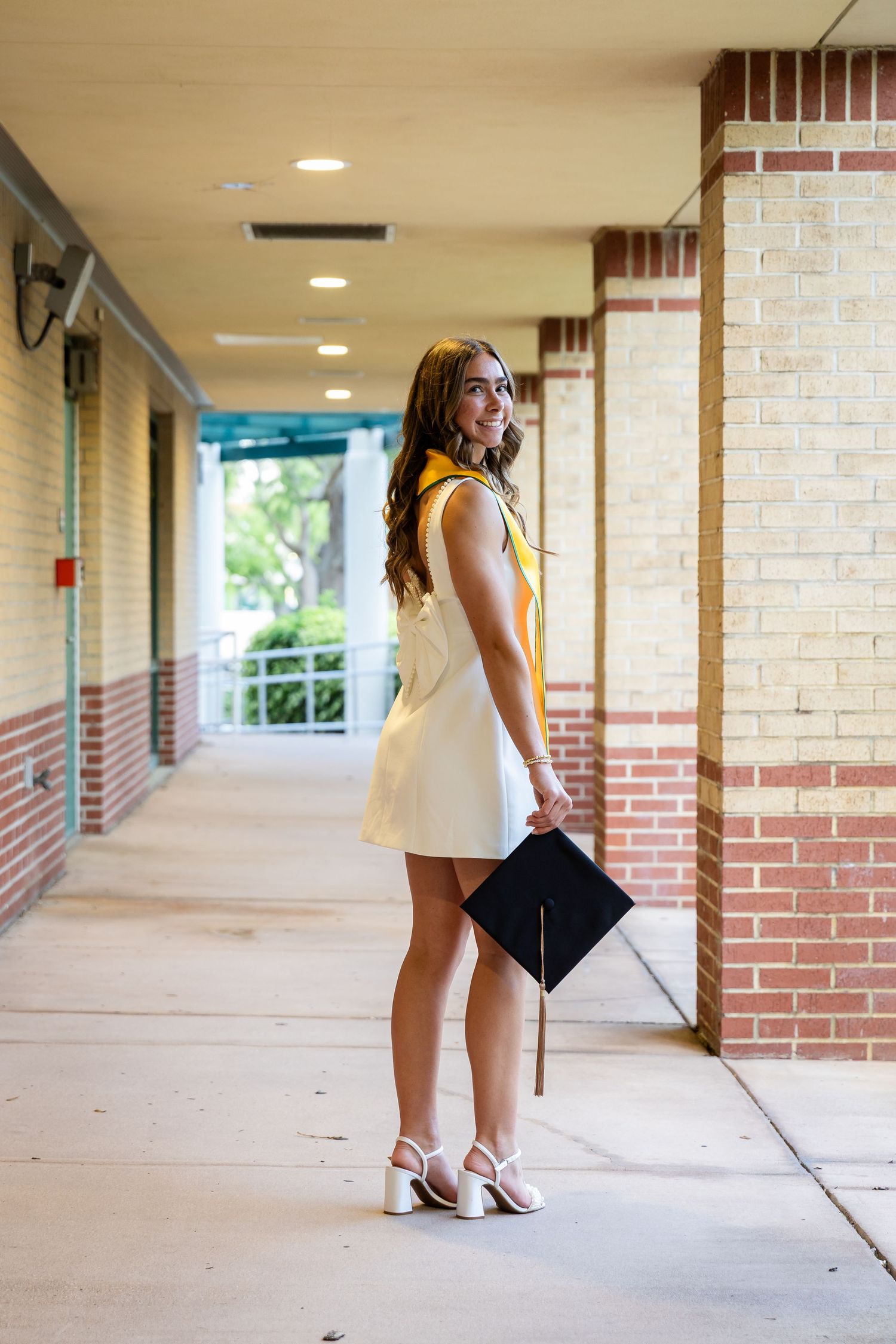 Young woman in white dress holding graduation cap smiles over shoulder in brick school hallway.