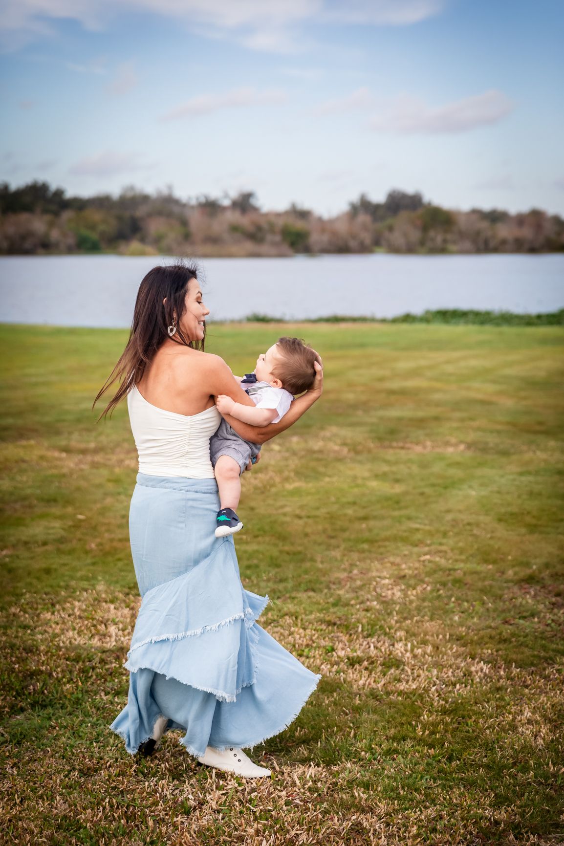 A mother in a white top and blue skirt joyfully holds her baby outdoors near a lake on a sunny day.