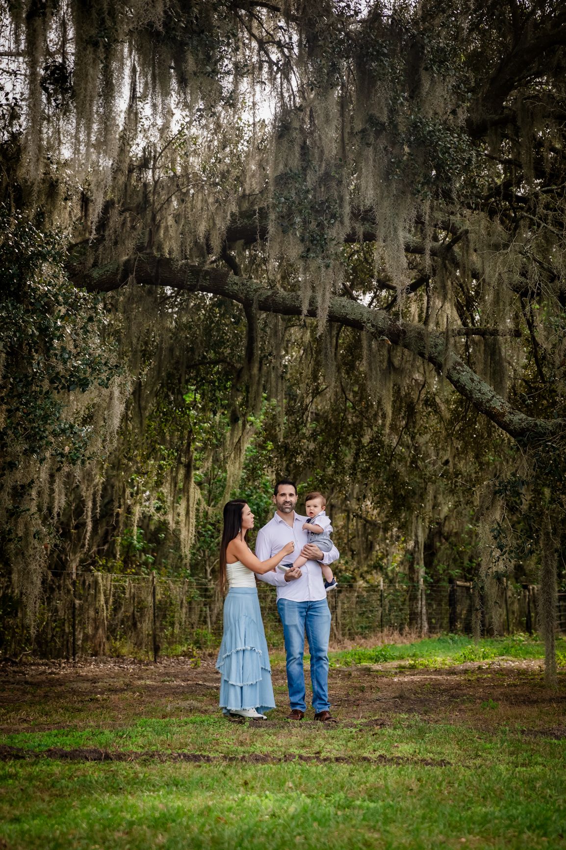 Young family standing under dramatic Spanish moss oak trees, father holding baby while mother gazes upward.