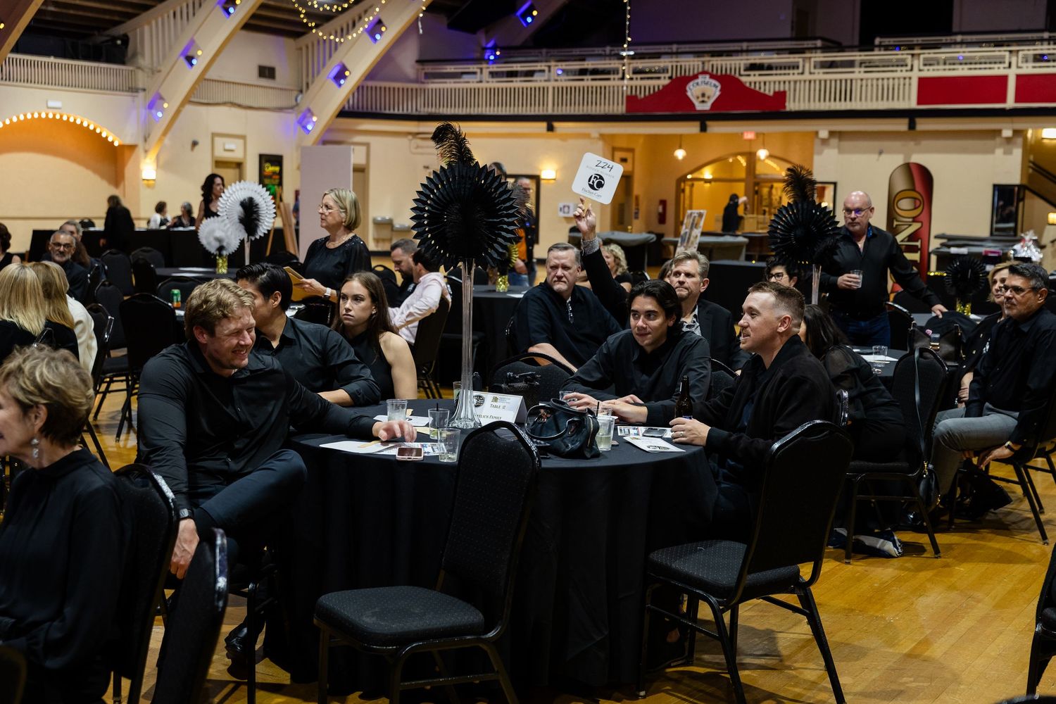 Group of people dressed in black seated at round tables during a formal event in a decorated ballroom.
