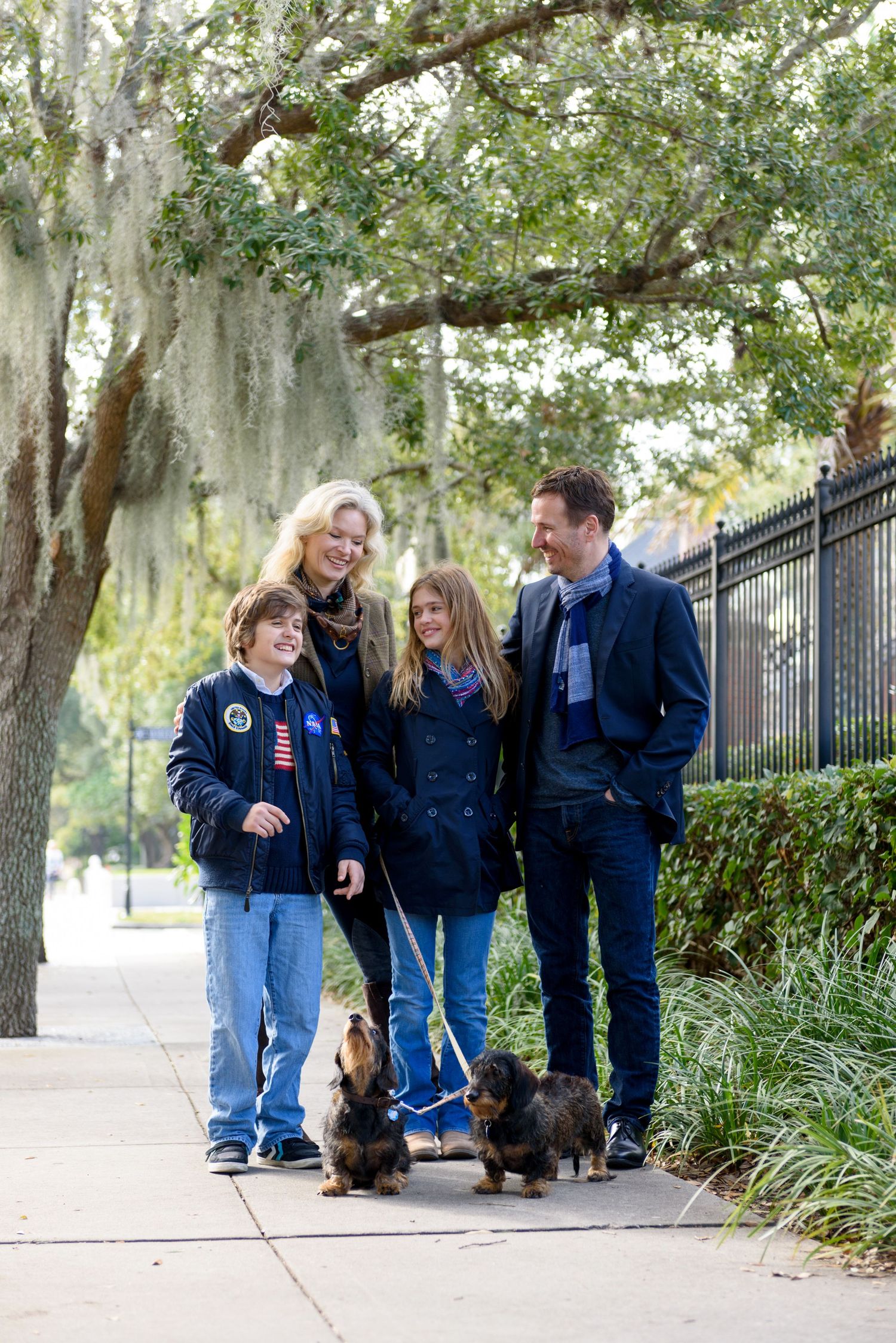 Family of four walking two dachshunds on a tree-lined sidewalk, smiling together on a sunny day.