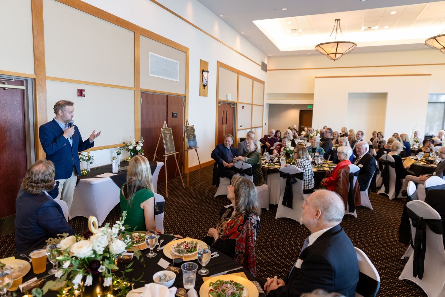 Man speaking at podium during formal banquet event with guests seated at decorated tables in elegant ballroom.