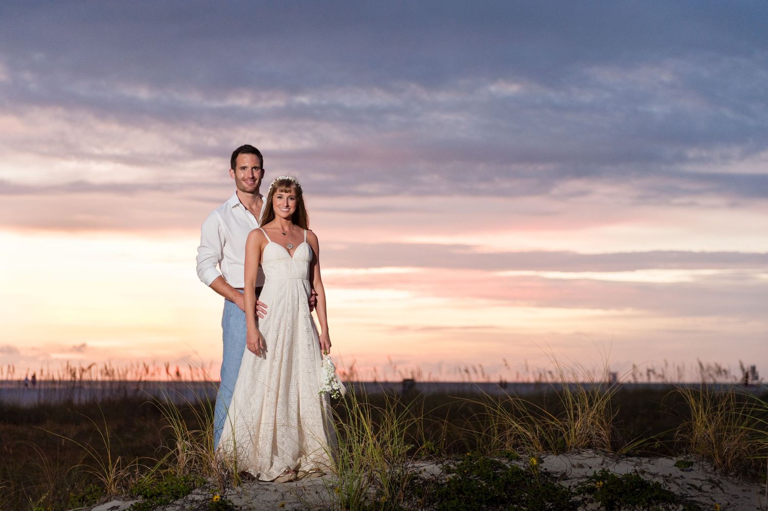 Couple in wedding attire poses on sandy beach dunes at sunset with colorful pink and purple sky backdrop.