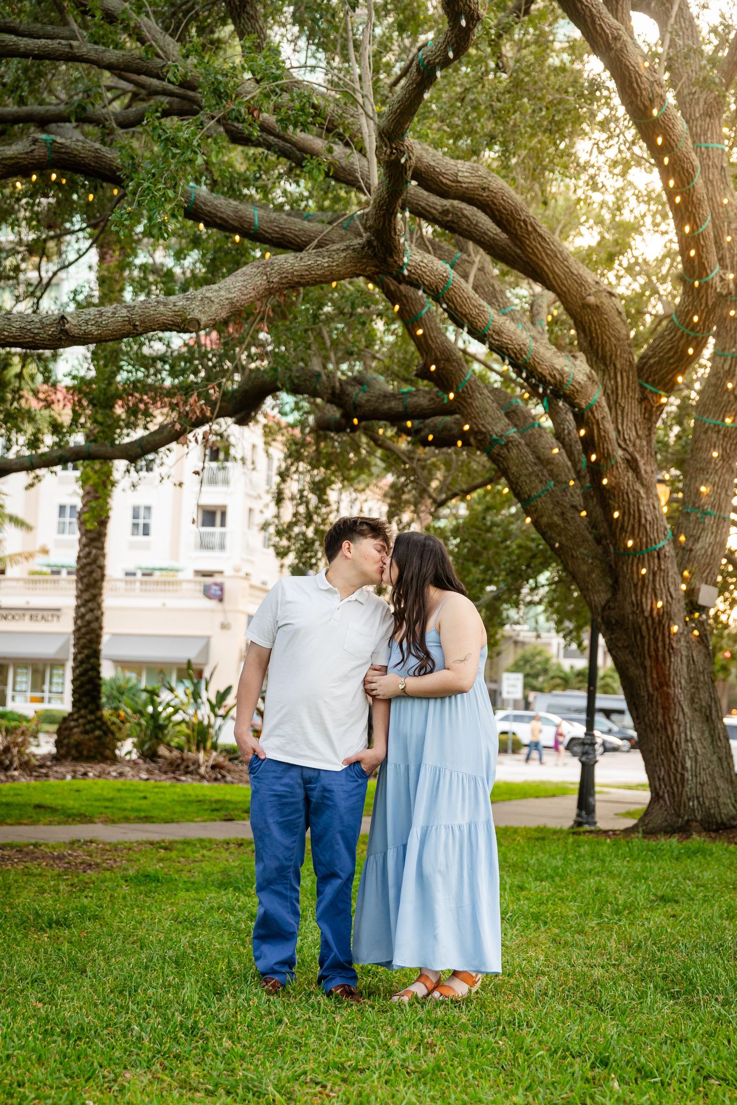 Couple sharing a kiss under large oak trees with string lights in a lush green park setting.