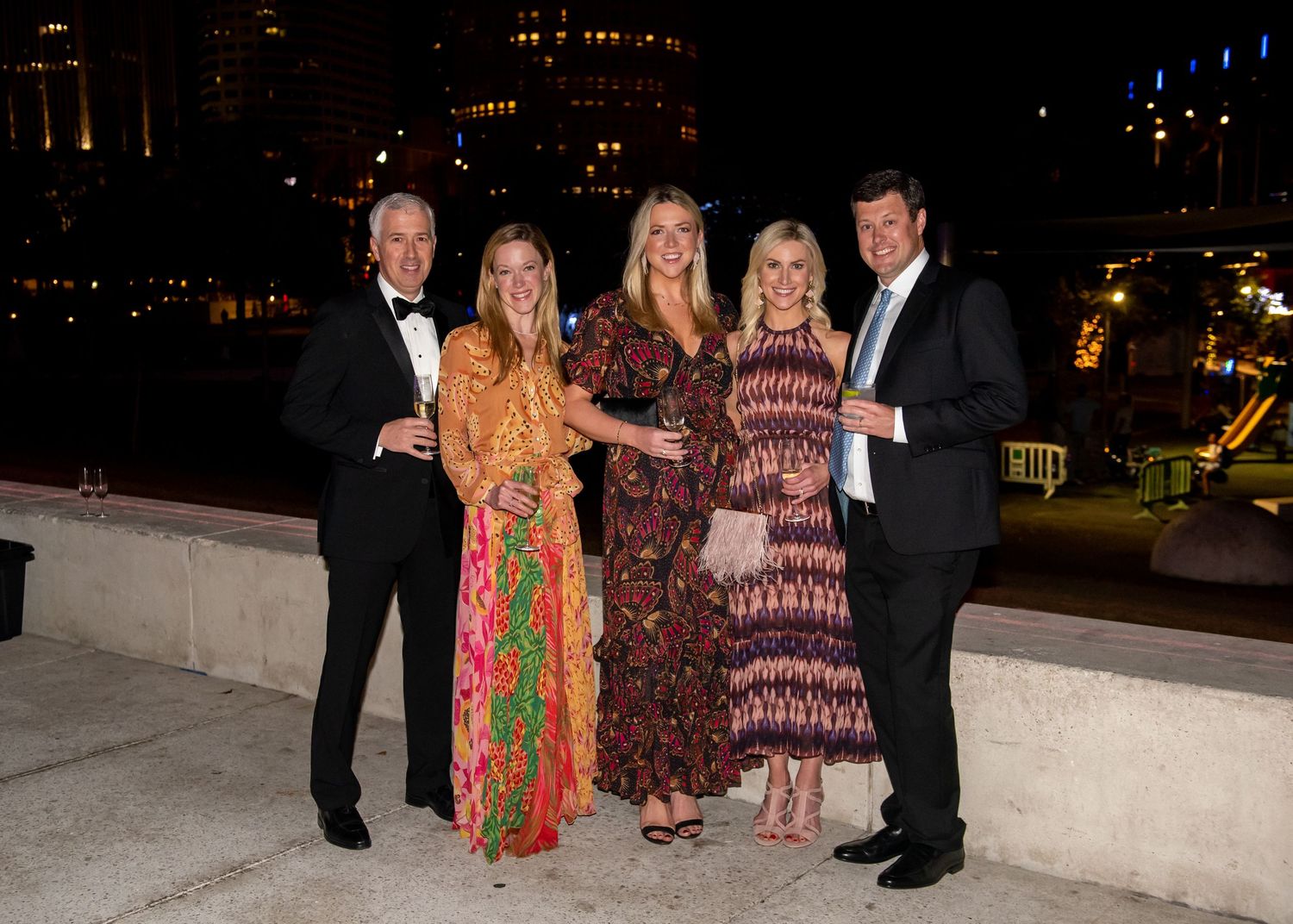 Five elegantly dressed guests pose together at an upscale evening outdoor event with city lights in the background.