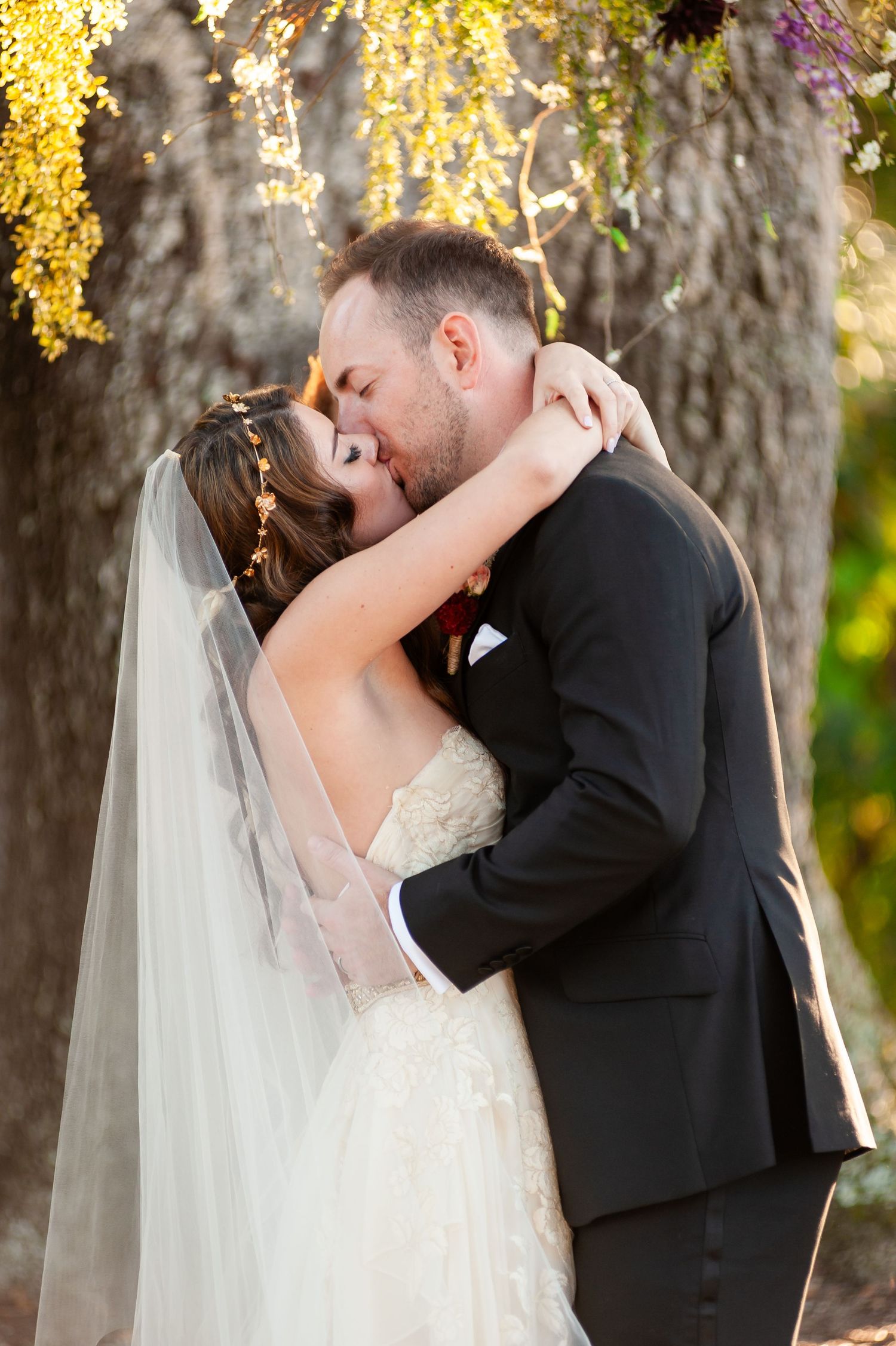 Newlywed couple shares first kiss under Spanish moss-draped trees with yellow wisteria arch backdrop.