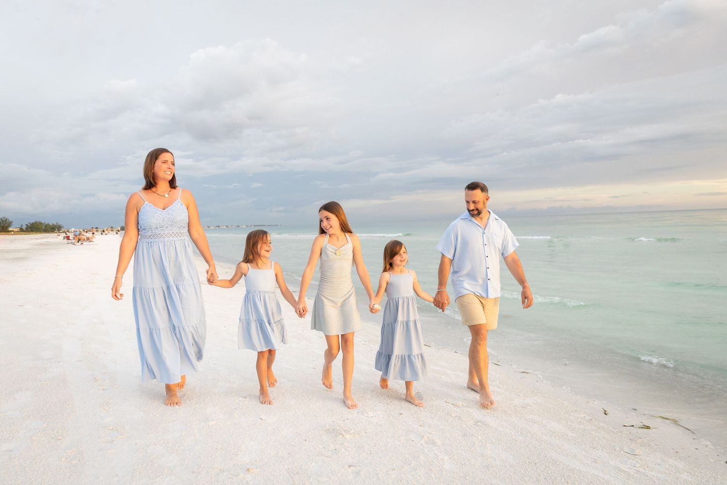Family of five walking hand-in-hand on a white sand beach, wearing coordinated blue and white outfits.