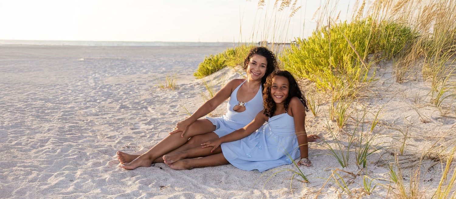 Mother and daughter sitting together on a sandy beach, wearing white dresses, with sea oats in the background.