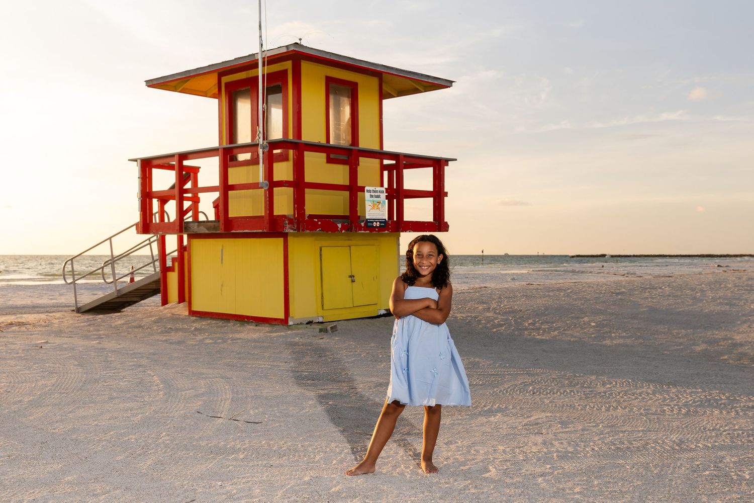 Young girl in blue dress stands barefoot on sandy beach beside colorful red and yellow lifeguard station at sunset.