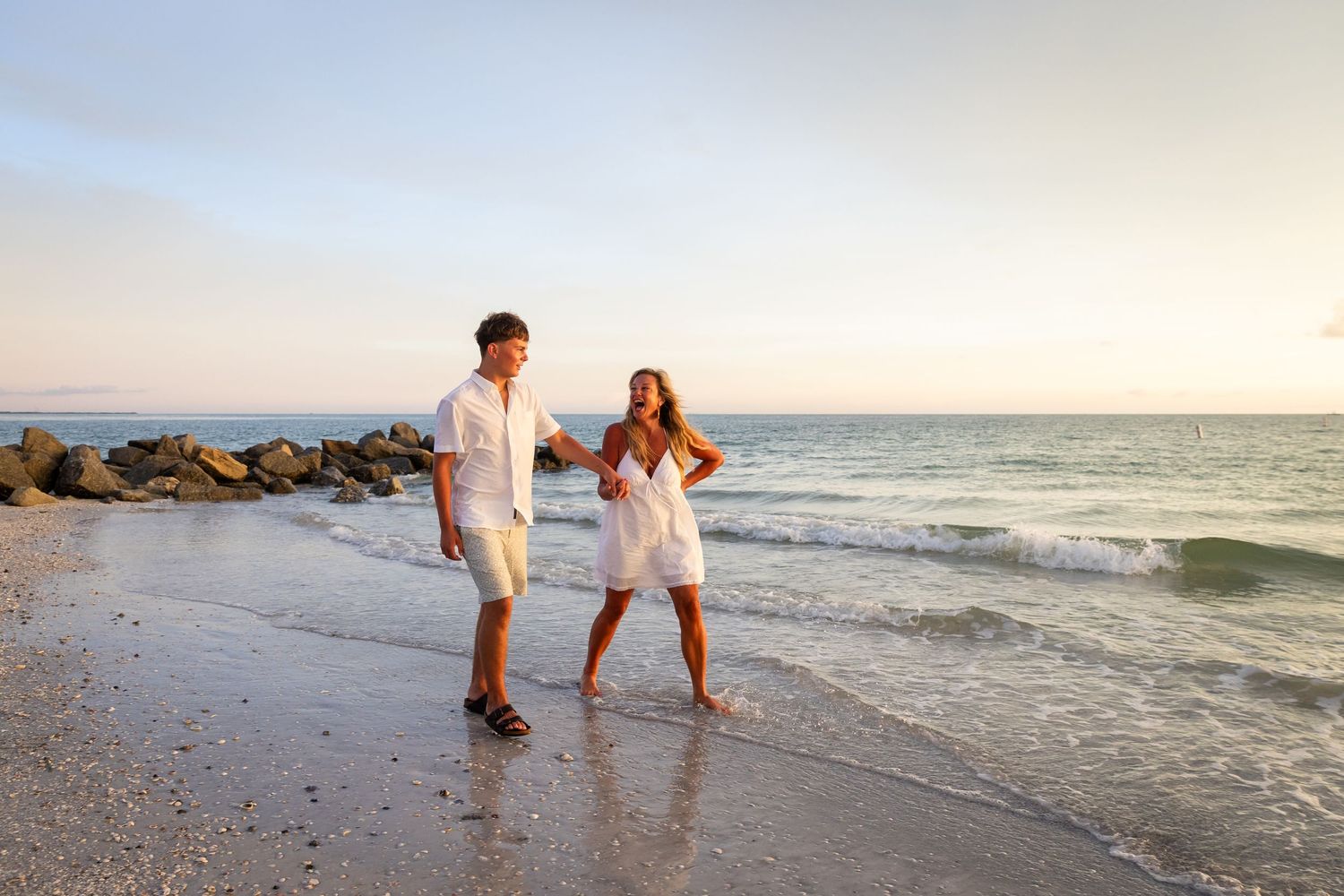 Couple in white outfits walking hand in hand on a sandy beach at sunset with gentle waves and rocky shore.