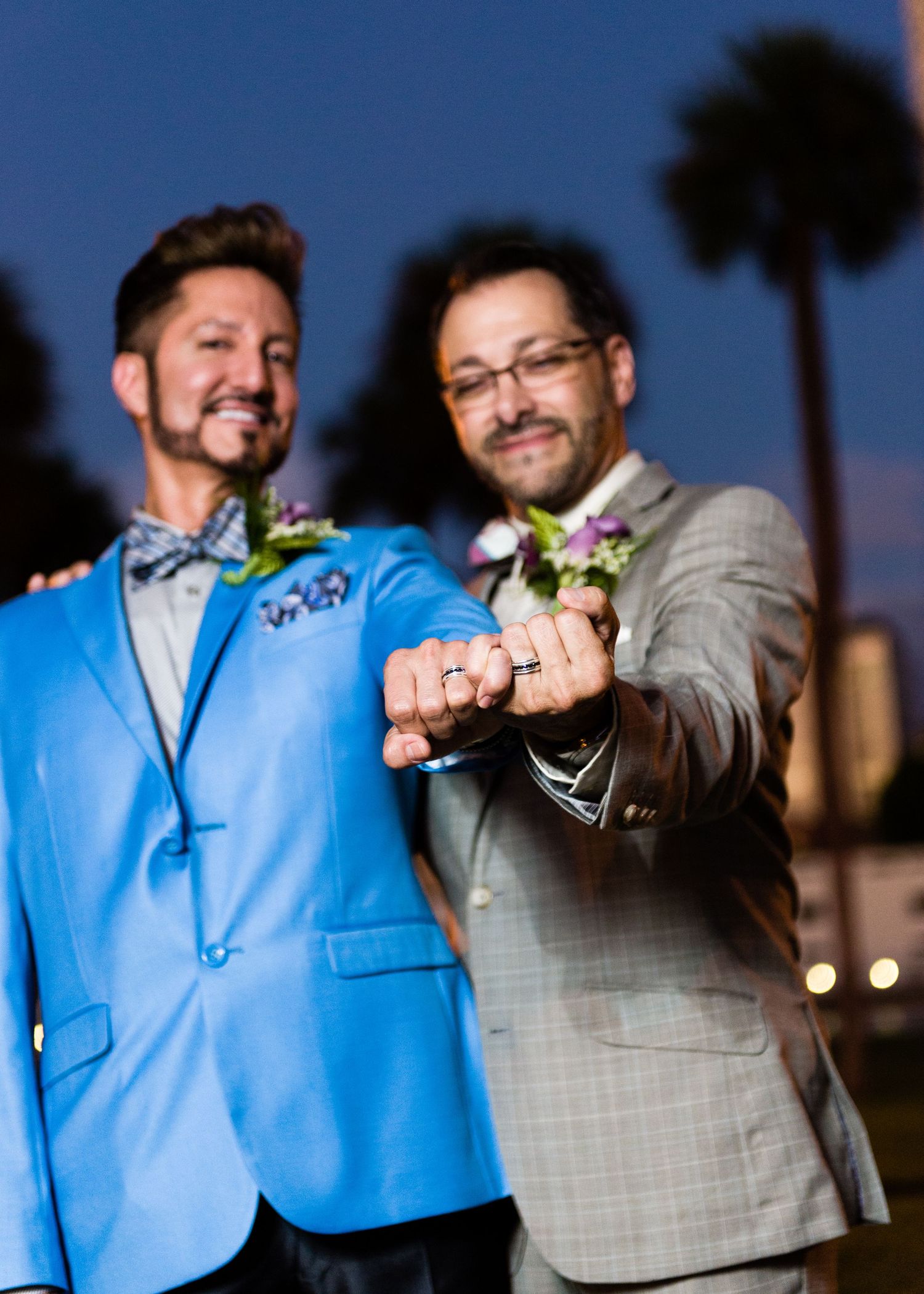 Two grooms in blue and gray suits show off wedding rings at dusk with palm trees in background.