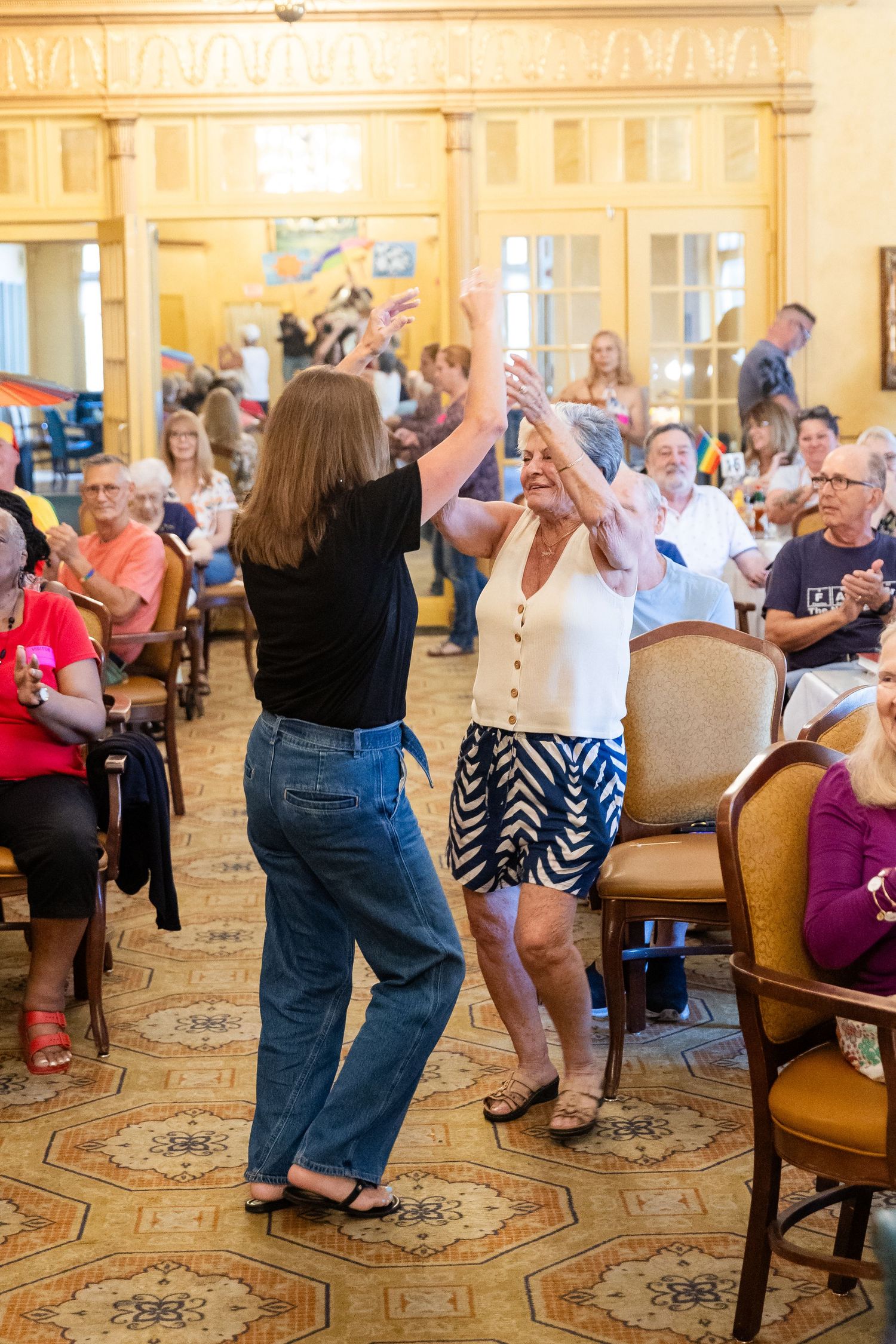Two women joyfully dancing with arms raised at a crowded indoor senior community gathering.