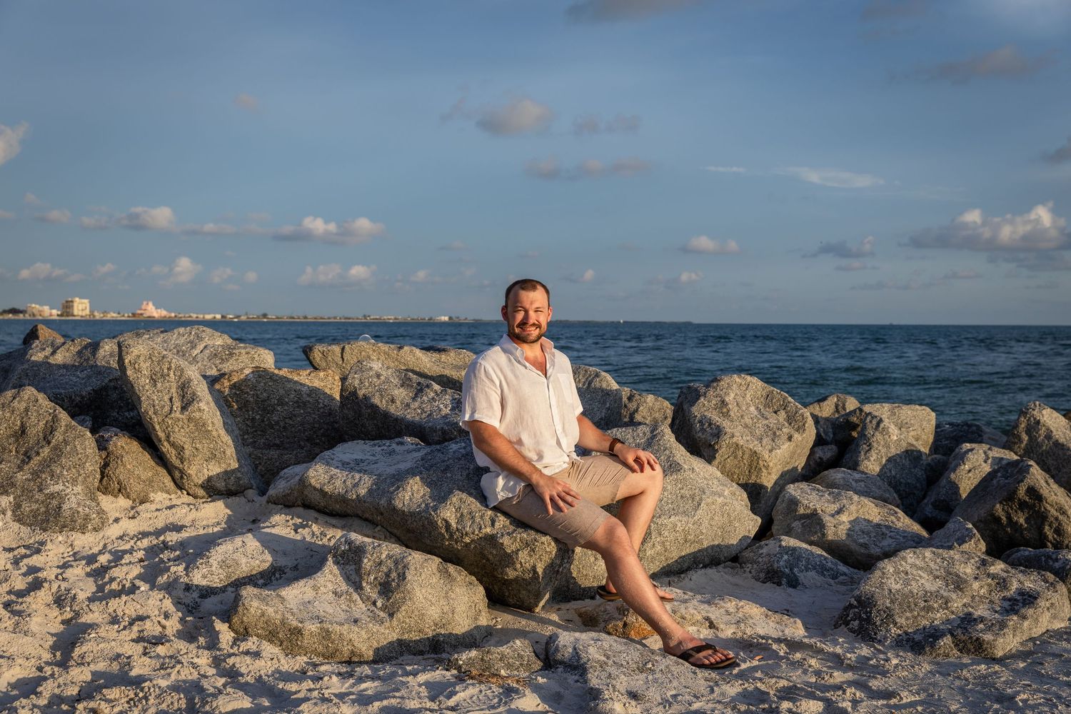 A man sits relaxed on rocky shoreline boulders by the ocean under a partly cloudy sky.