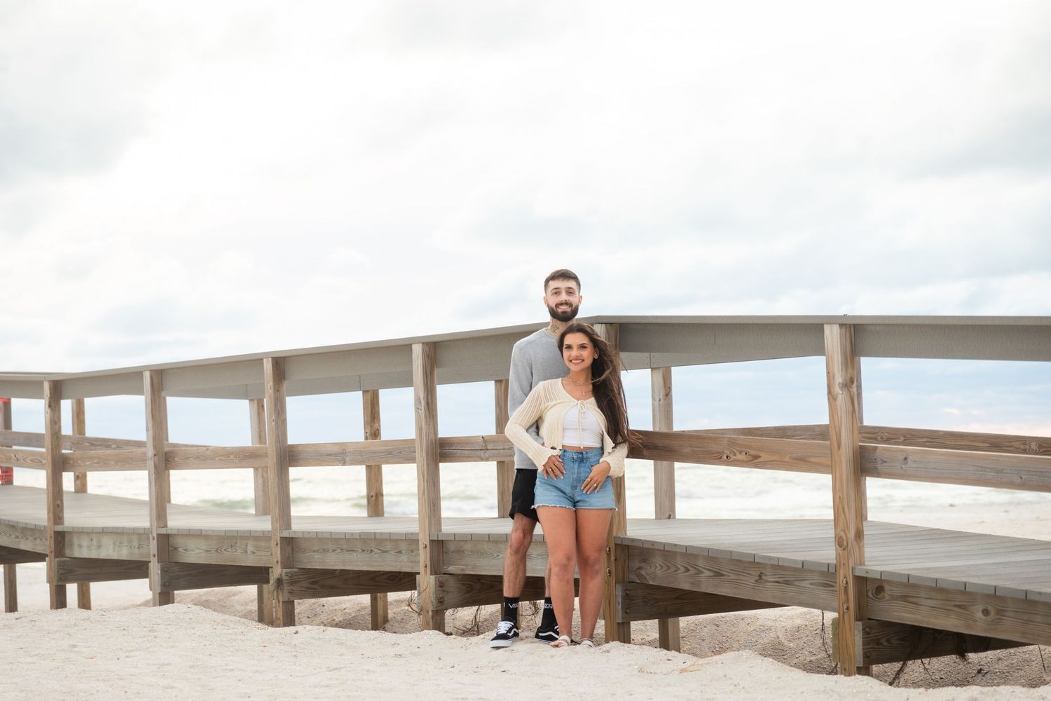 Couple posing together on a wooden beach boardwalk with sandy shores and cloudy sky in the background.