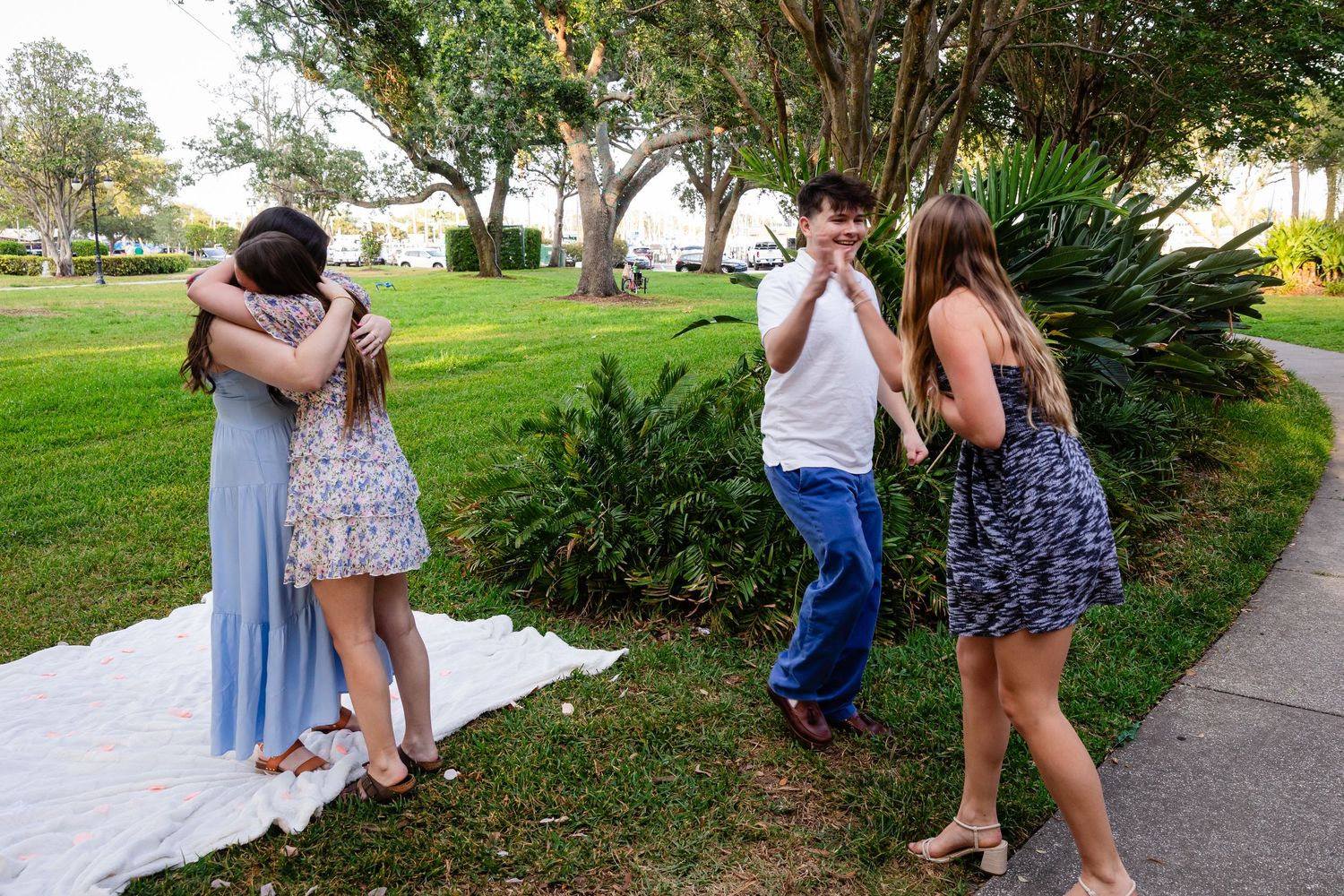 Four young people celebrate outdoors on a sunny day, two hugging warmly while others laugh and chat on a green lawn.