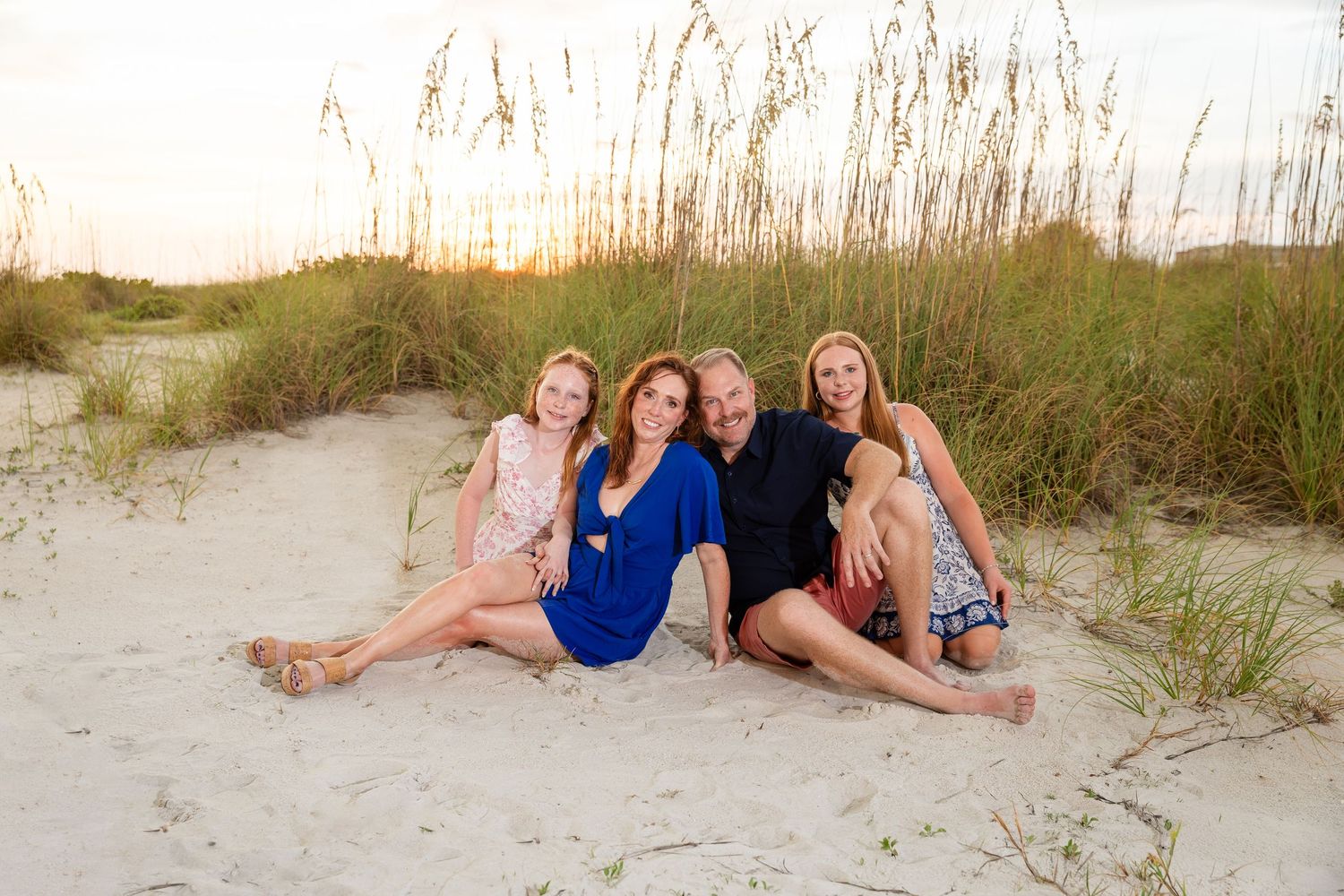 Family of four smiling together on sandy beach dunes at sunset with sea oats in background.