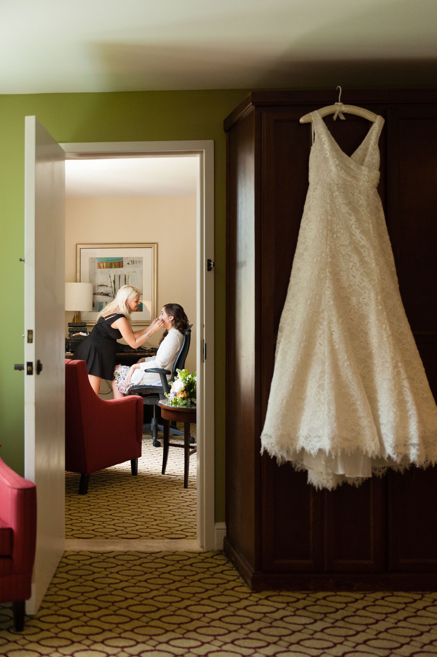 White lace wedding dress hanging on wooden wardrobe while bride gets makeup done in hotel suite background.