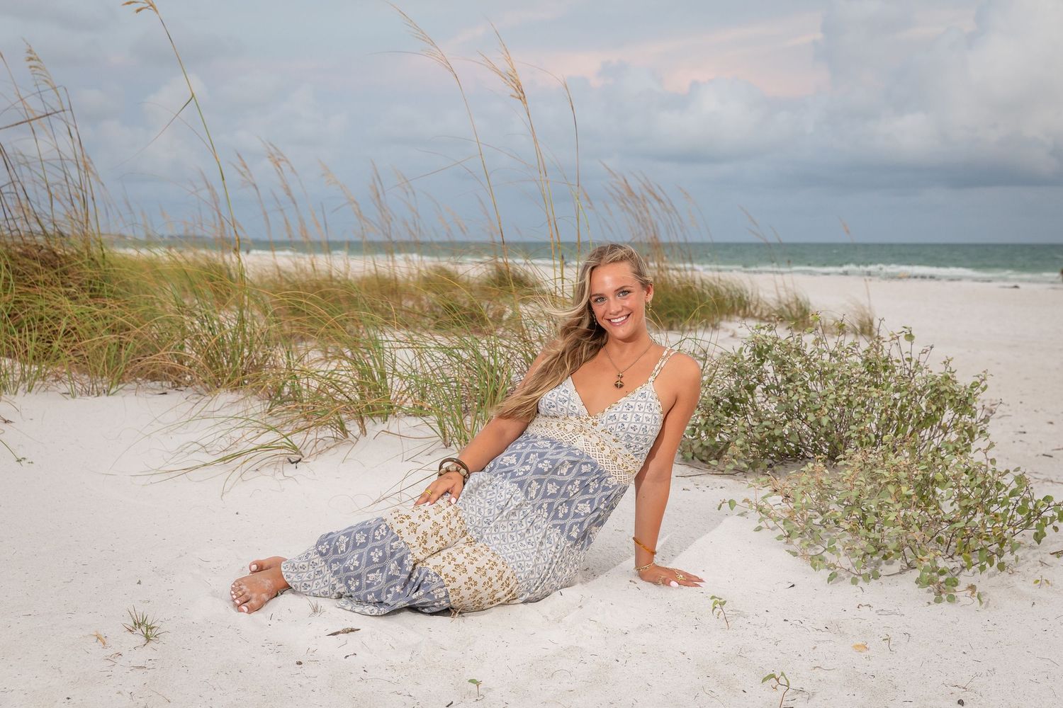 Young woman in floral maxi dress posing on white sand beach dunes with sea oats and ocean in background.