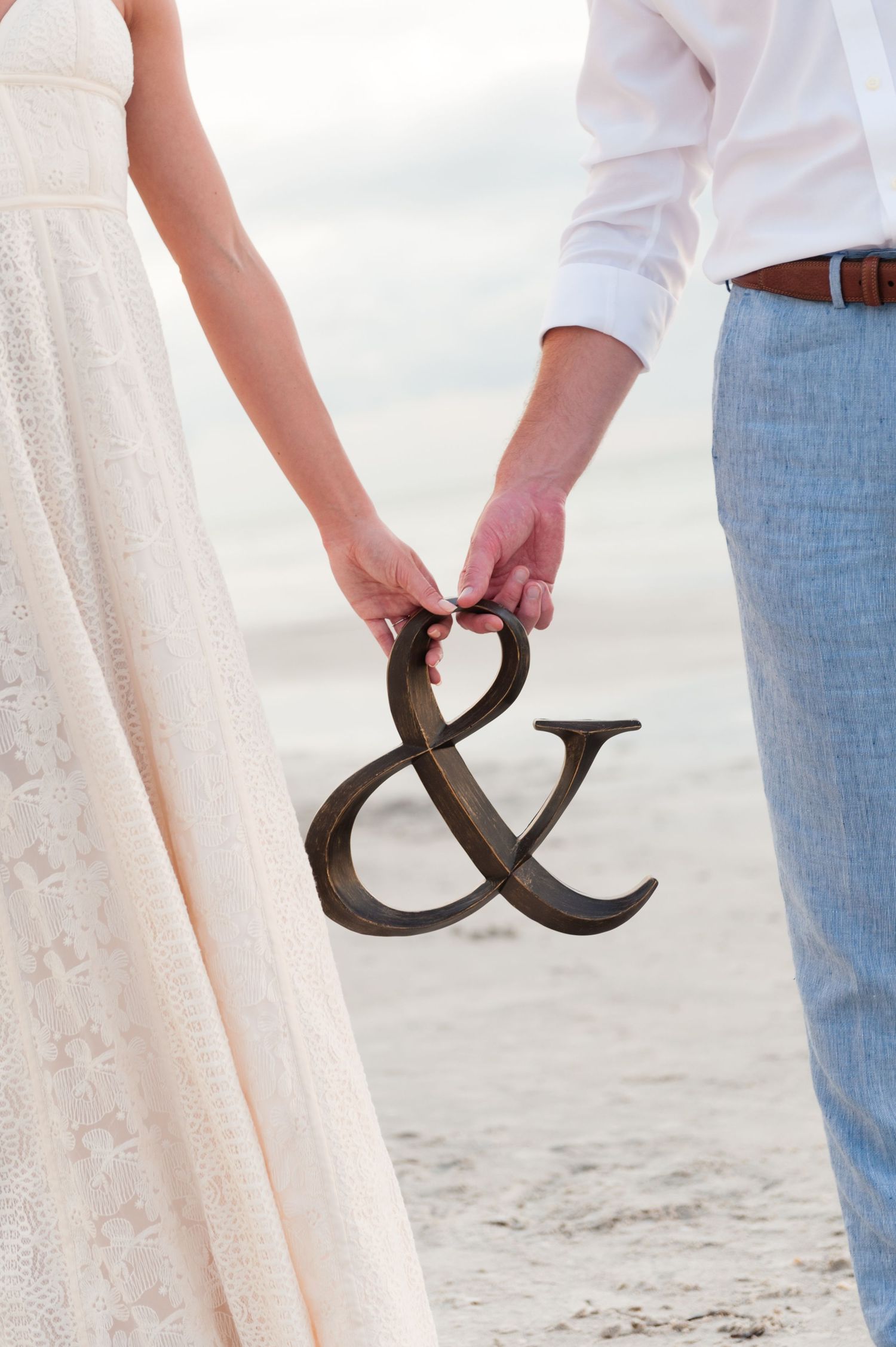 Bride and groom holding a rustic ampersand symbol together on the beach after wedding.