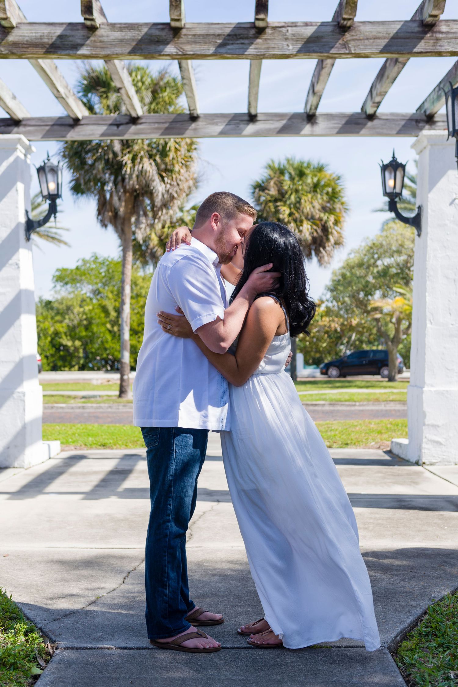 Couple sharing a romantic kiss under a white pergola with palm trees in the background.