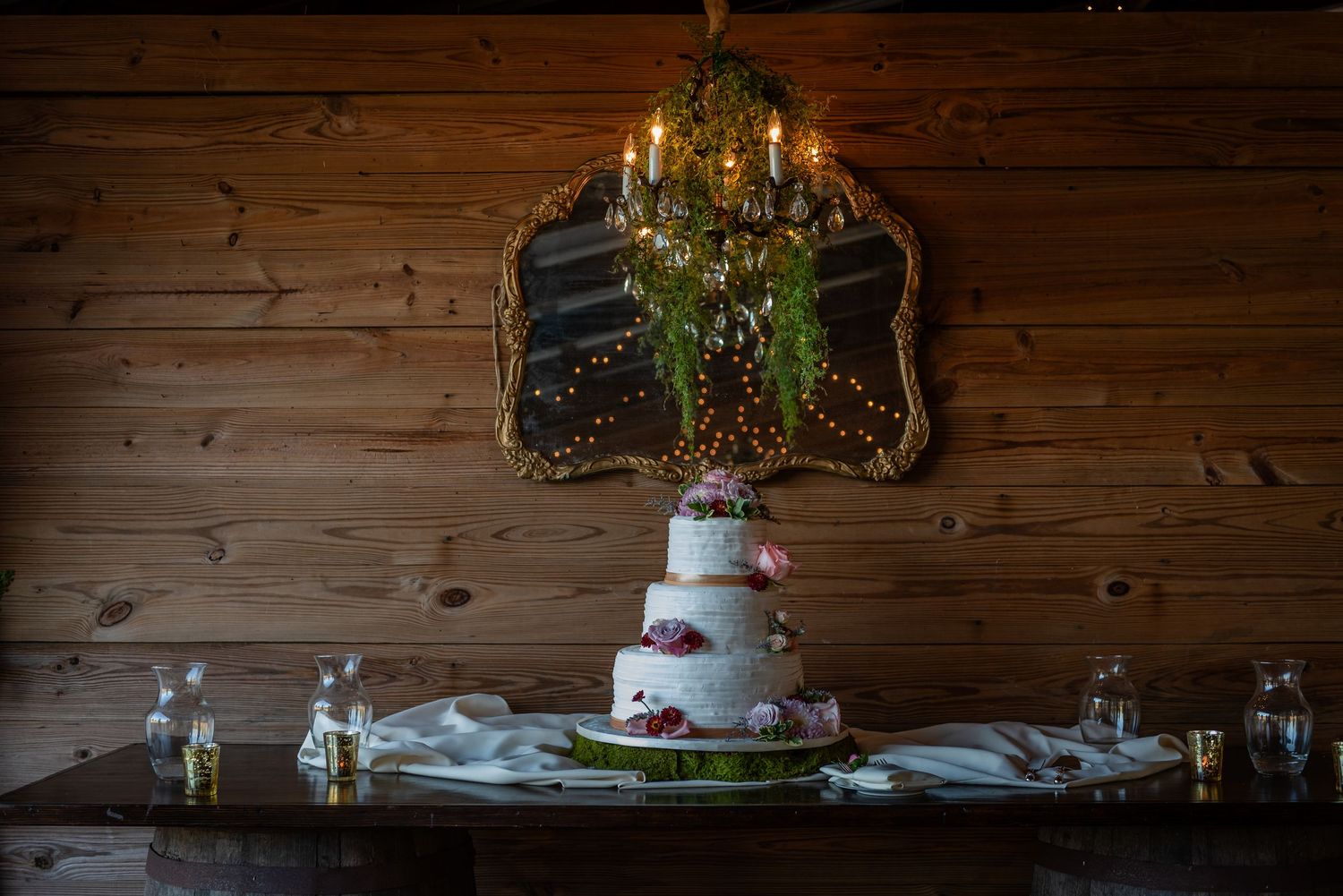 Elegant three-tier white wedding cake displayed on a wooden table beneath a chandelier with string lights.