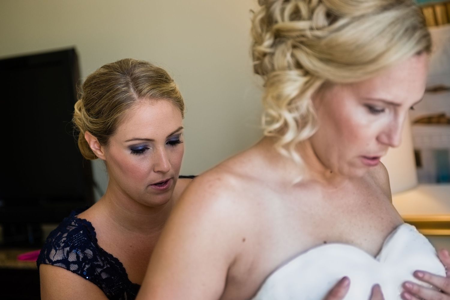 Bridesmaid in navy lace dress helping blonde bride get dressed before wedding ceremony.
