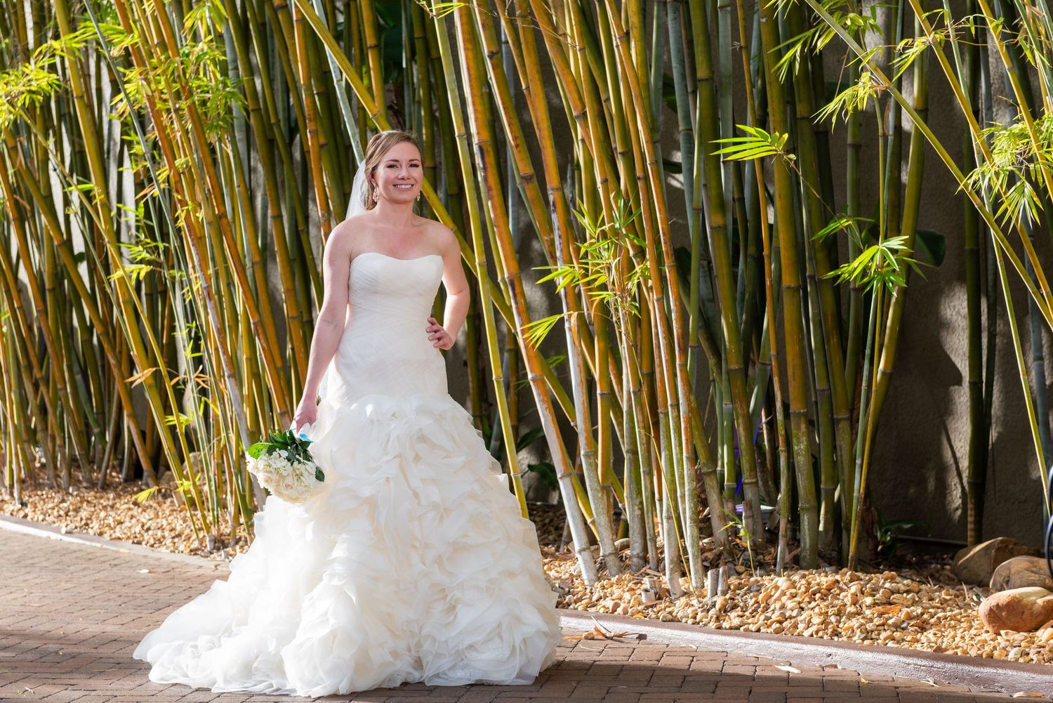 Bride in strapless ruffled white wedding gown holding bouquet poses before tall bamboo plants outdoors.