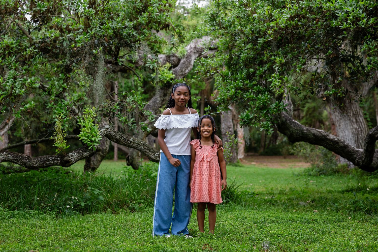 Two girls pose together on green grass surrounded by lush trees and rocky landscape in an outdoor portrait.