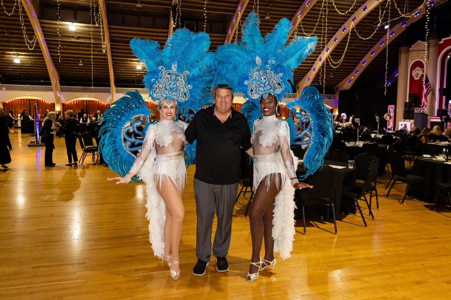 Man poses with two showgirls wearing elaborate blue feathered headdresses at a Las Vegas-style gala event.