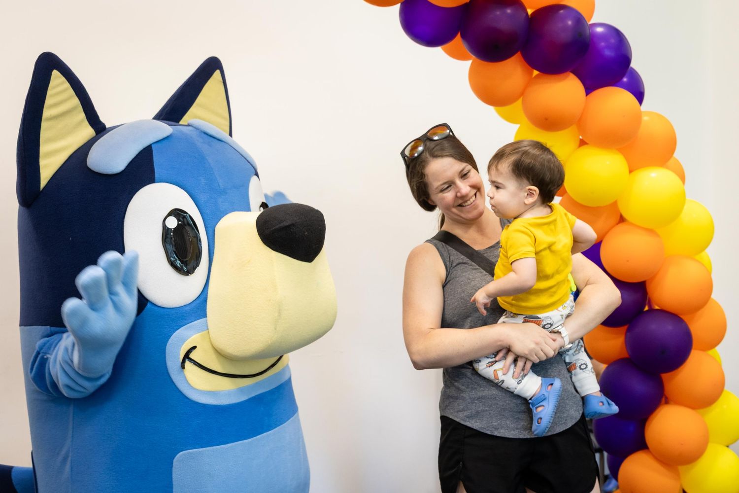 Woman holding baby smiles next to Bluey mascot costume character with colorful balloon arch backdrop.