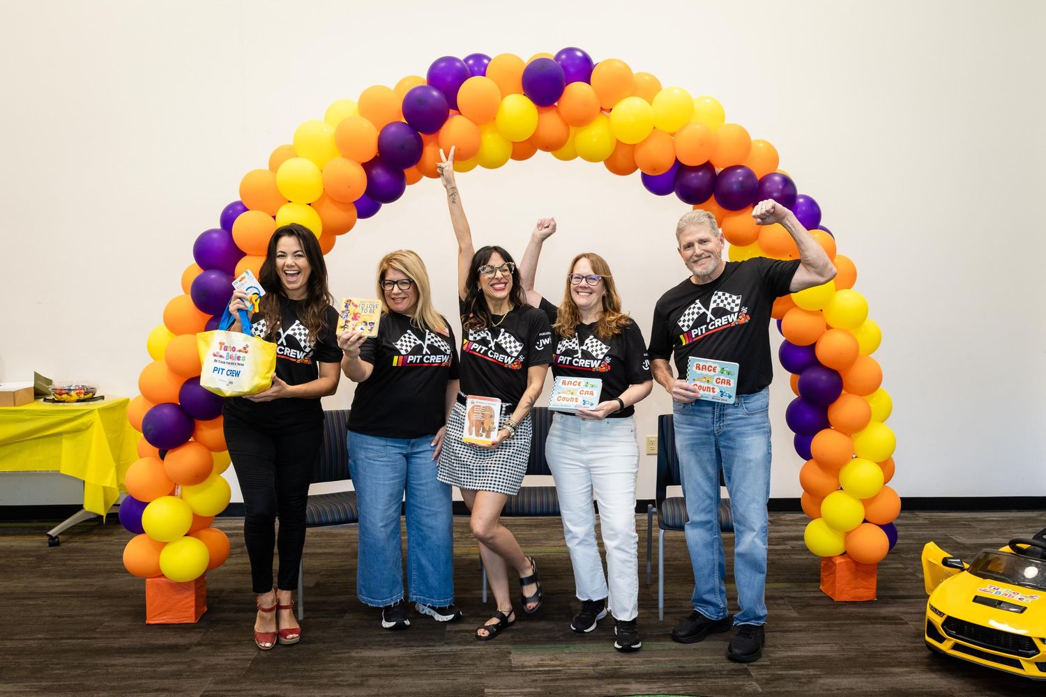 Five people pose under a colorful orange and purple balloon arch, holding awards and celebrating at an indoor event.