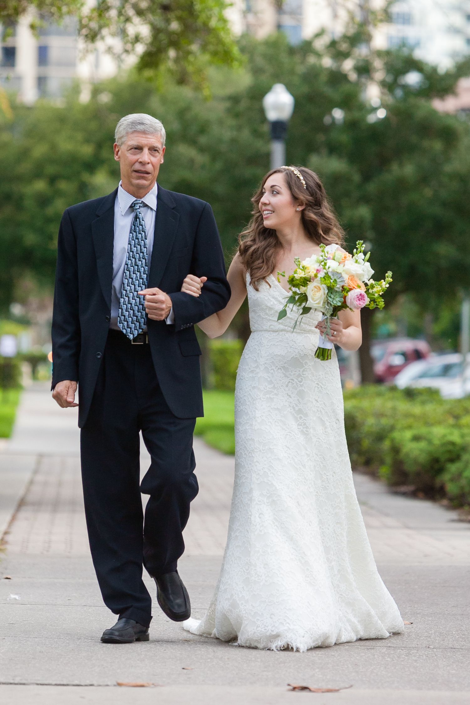 Bride in white lace gown holds pastel bouquet while walking with father in navy suit on outdoor path.