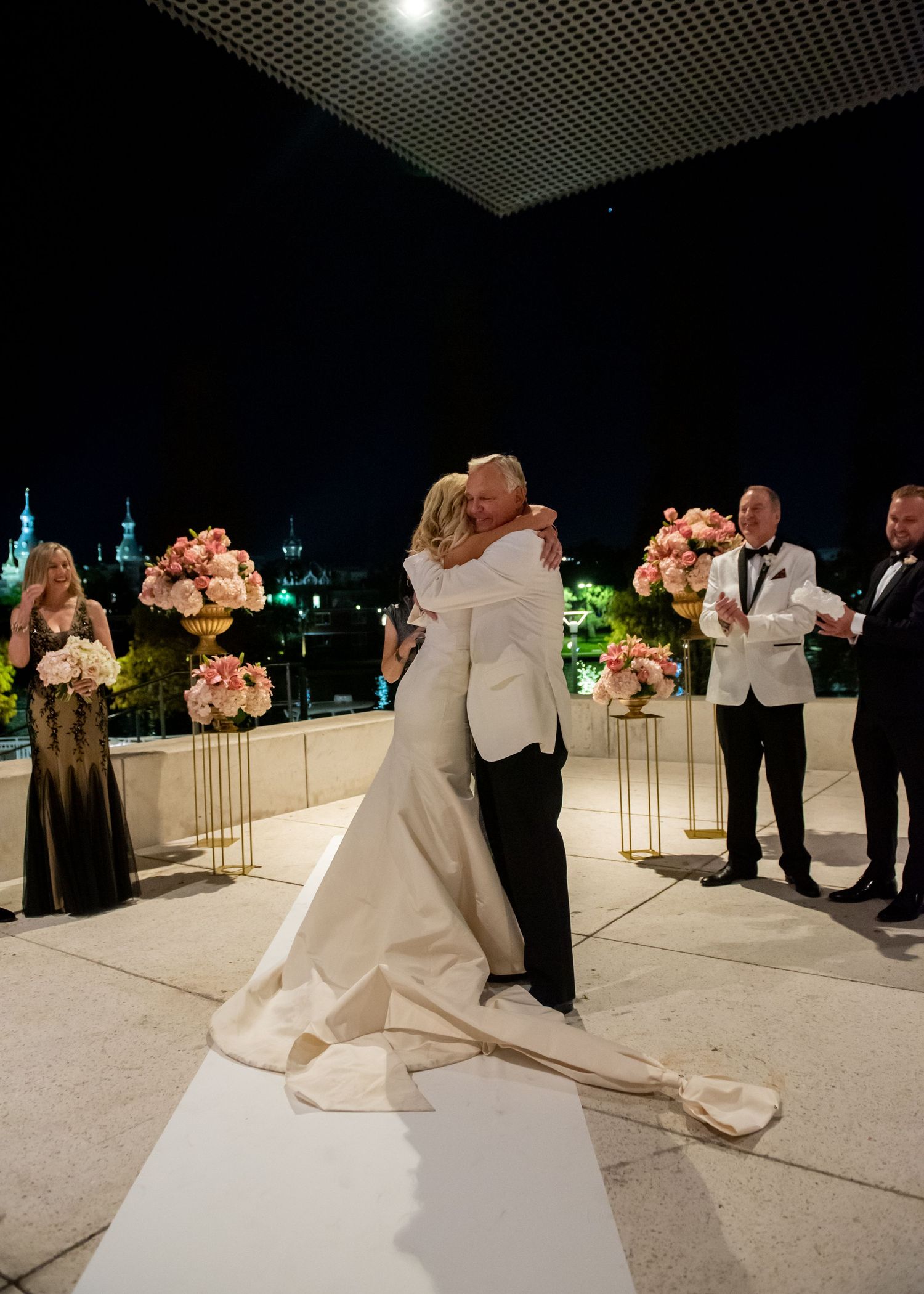 Bride and groom share first kiss at outdoor rooftop wedding ceremony at night with pink floral arrangements.