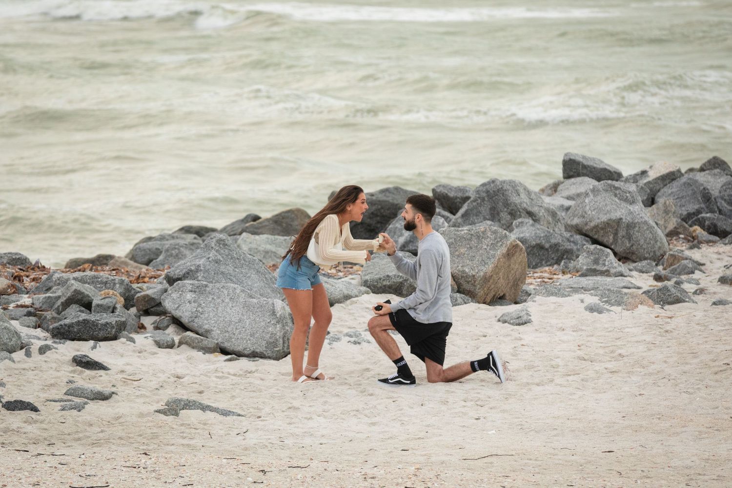 Man proposes to woman on a sandy beach beside large rocks with ocean waves in the background.