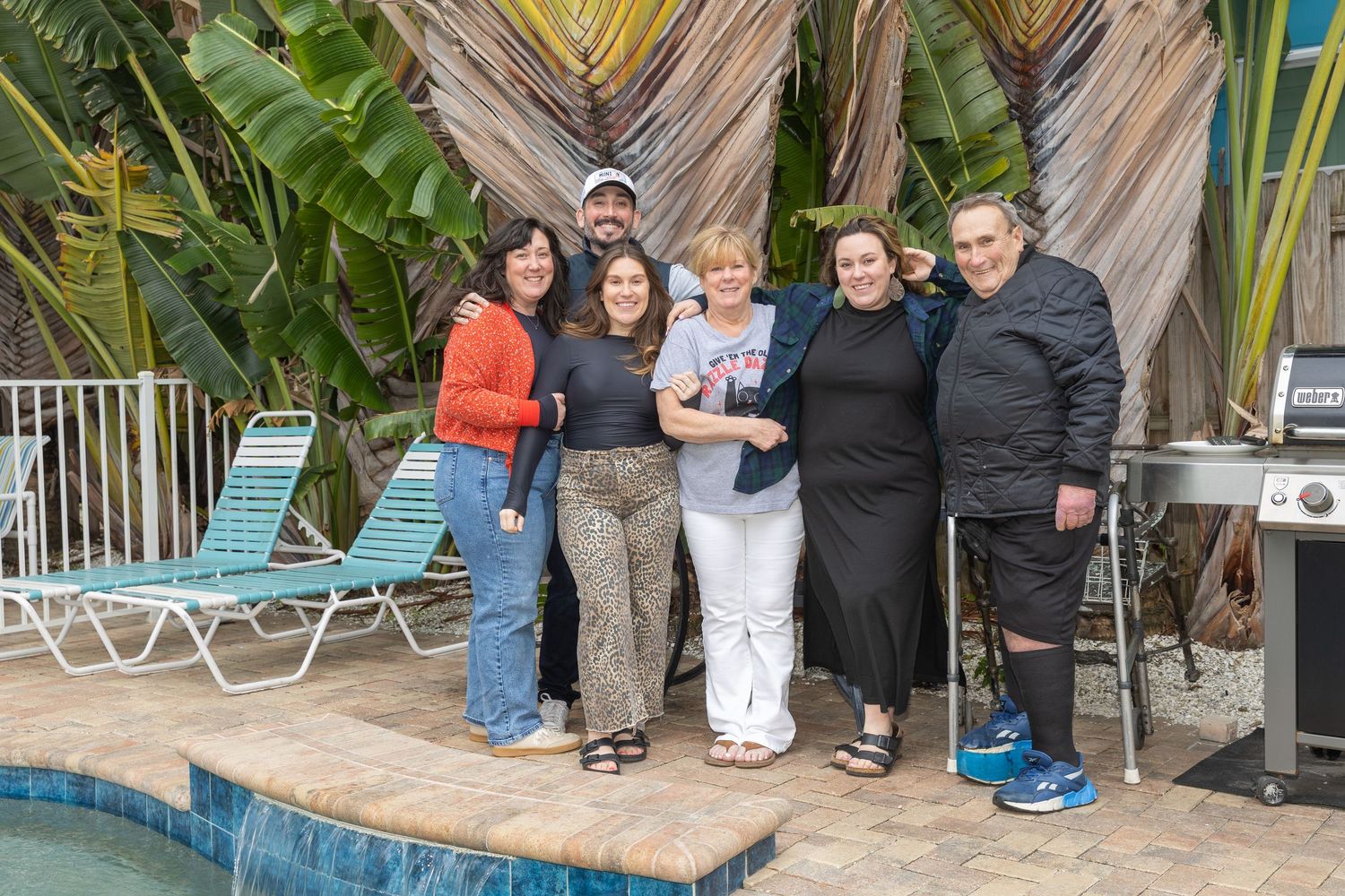 Group of six people smiling together outdoors near a pool surrounded by tropical plants.