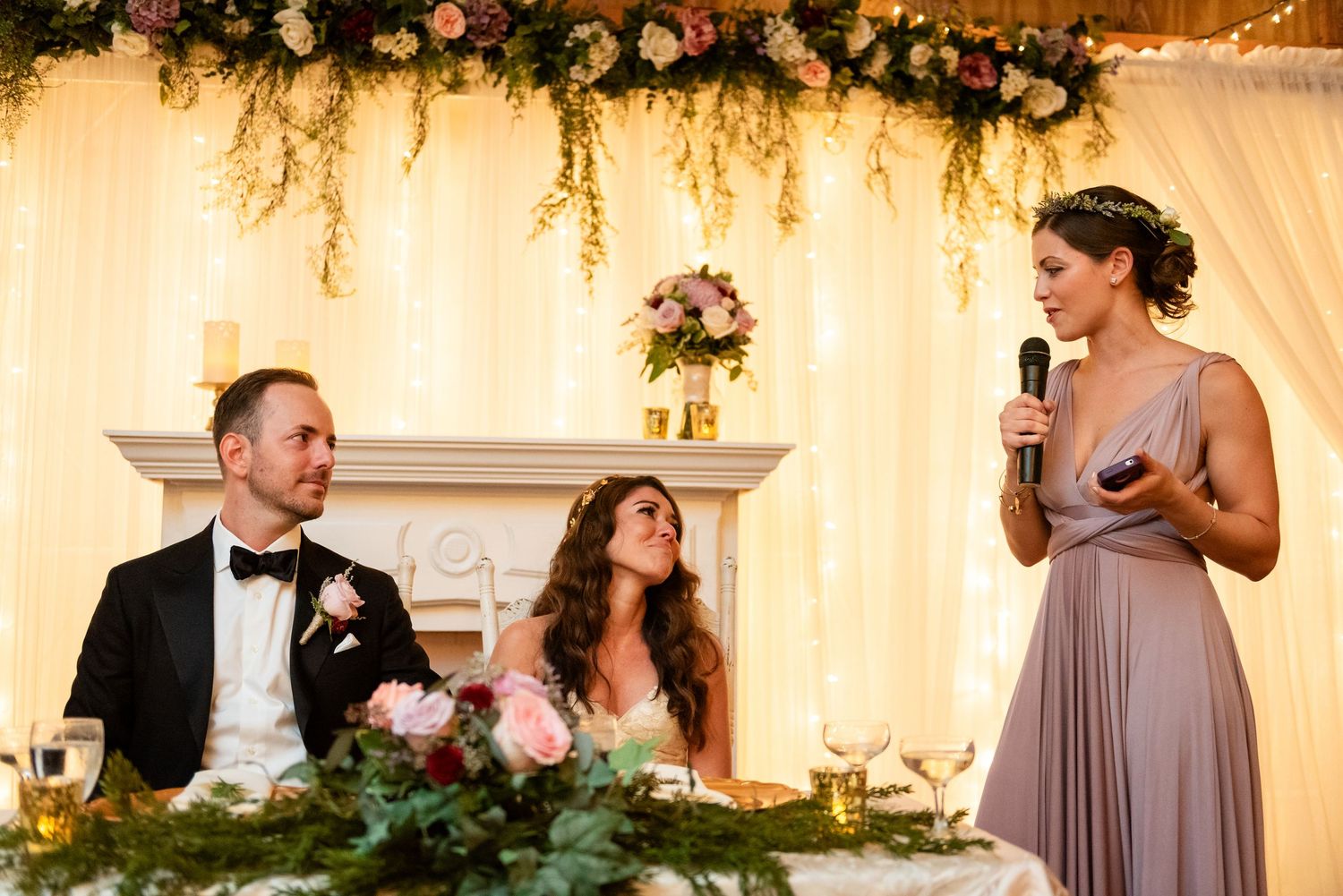 Bridesmaid in mauve dress giving speech at wedding reception while bride and groom listen at head table.