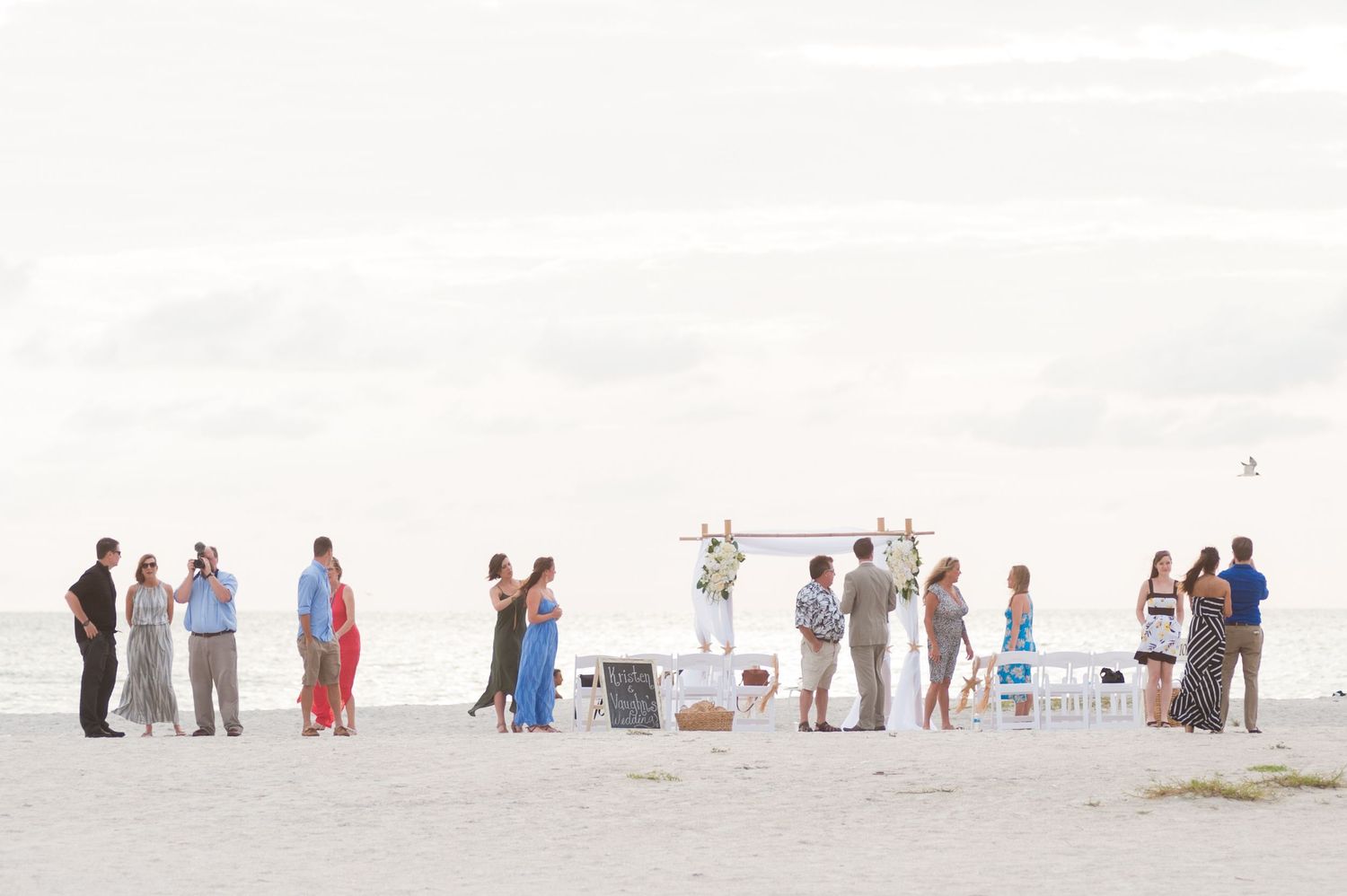 Small beach wedding ceremony with guests gathered around a floral arch on white sand with calm ocean backdrop.