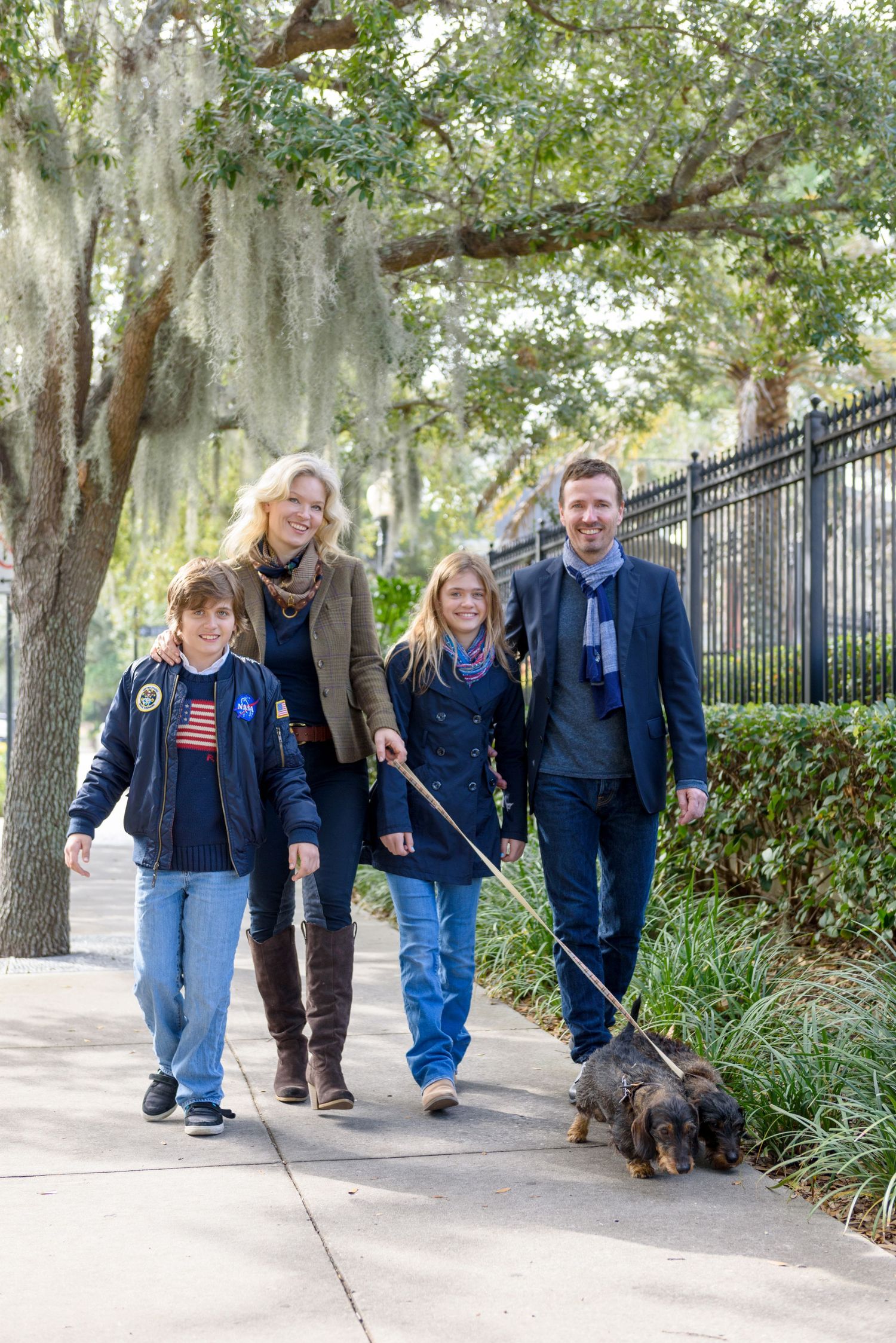 Family of four walking a dachshund on a tree-lined sidewalk on a sunny day.